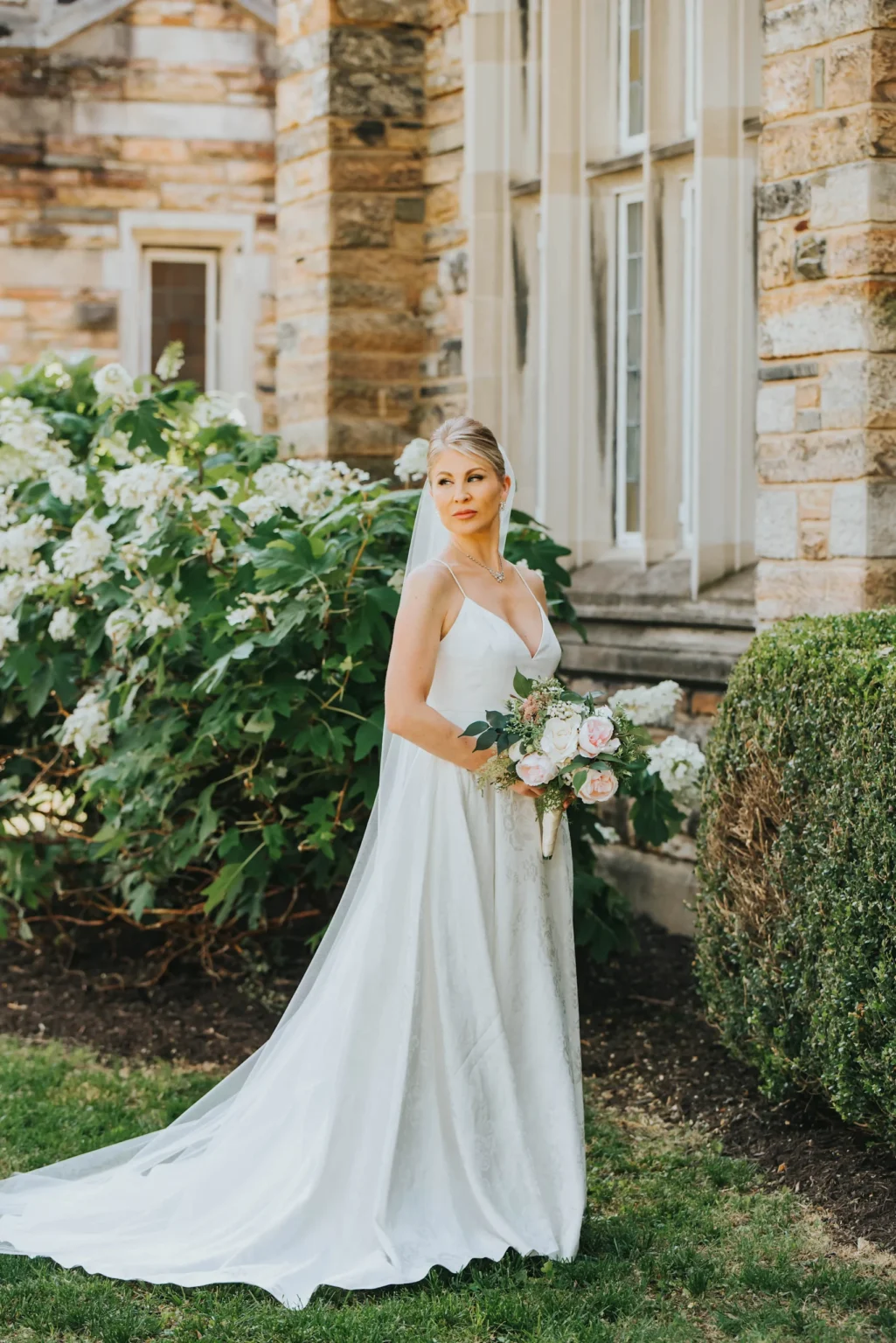 Elopement Wedding A bride stands outdoors near a stone building, facing slightly to the left and holding a bouquet of pink and white flowers. She is wearing a long white dress and a veil. Behind her is a lush green bush with white flowers. The scene is bright and serene, with natural sunlight illuminating the setting. Elopements Inc