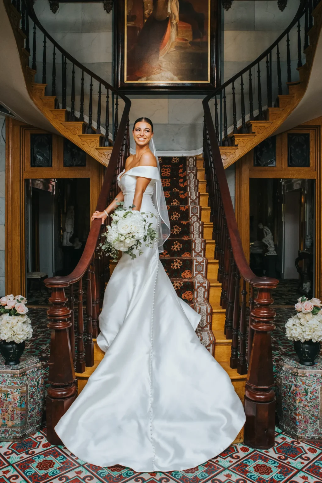 Elopement Wedding A bride stands gracefully in a satin off-the-shoulder wedding gown with a long train on a grand staircase. She holds a bouquet of white flowers and wears a veil. The staircase is ornate with wooden bannisters, floral arrangements, and a large framed painting hangs at the top. Elopements Inc