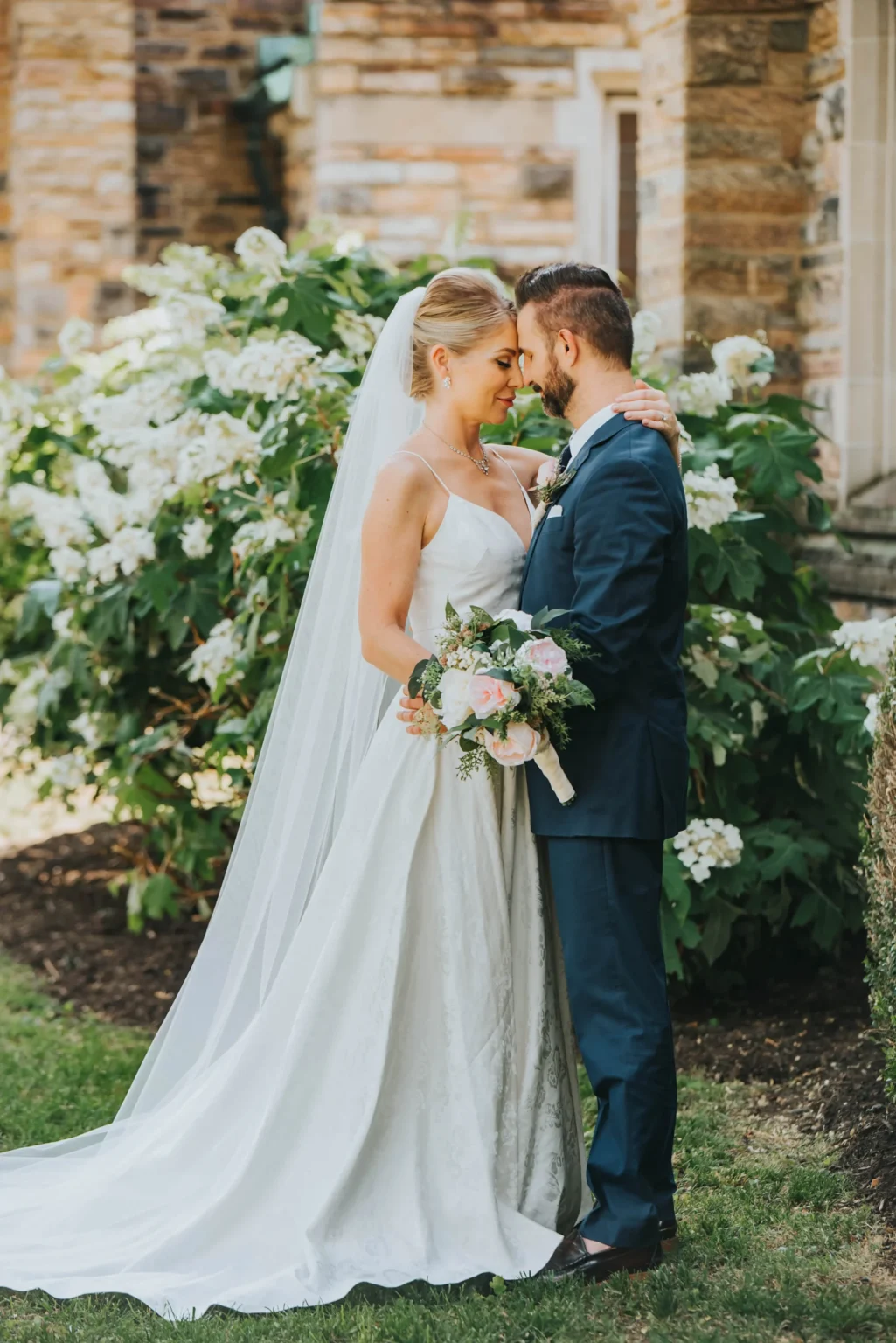 Elopement Wedding A bride in a white gown and veil, holding a bouquet of flowers, stands close to a groom in a dark blue suit. They are touching foreheads and embracing in front of lush green bushes with white flowers, beside a stone wall in an outdoor setting, possibly a garden or park. Elopements Inc