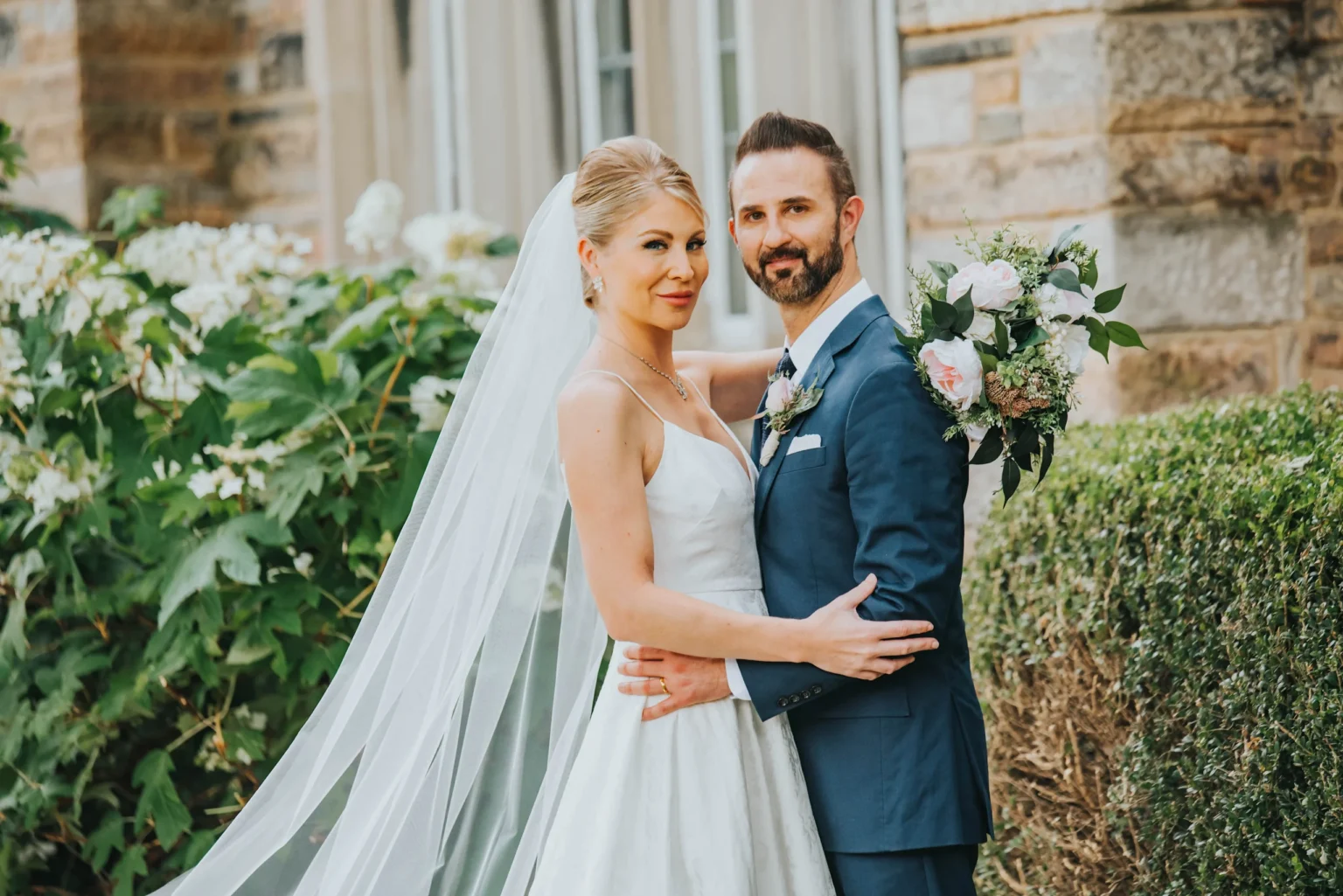 Elopement Wedding A bride and groom pose outdoors on their wedding day. The bride wears a white dress with a long veil, and has blonde hair styled up. The groom is in a navy blue suit with a white boutonnière. They stand close, with greenery and a brick building in the background. Both are smiling. Elopements Inc