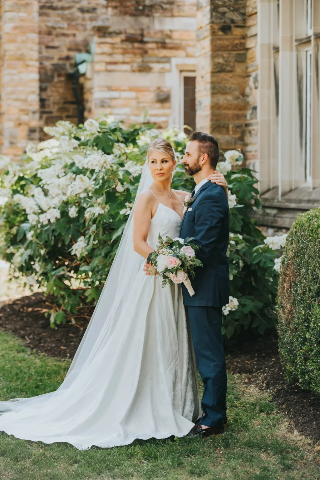 Elopement Wedding A bride in a white gown and veil stands close to a groom in a navy suit and white shirt. They are outdoors near a stone building with green foliage and white flowers in the background. The bride holds a bouquet with pink and white flowers as both look into the distance. Elopements Inc