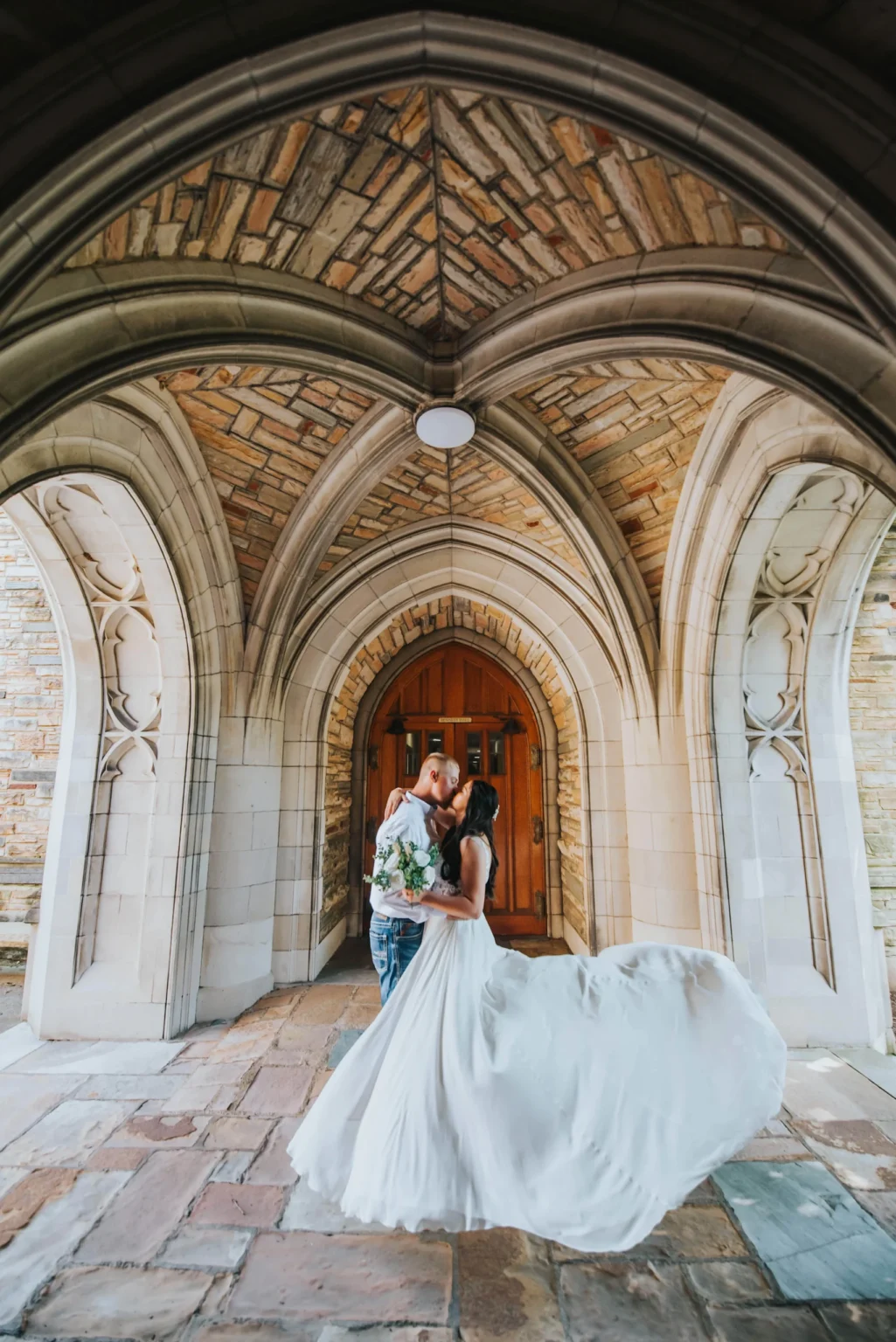 Elopement Wedding A couple shares a kiss under a stone archway. The groom, in a light blue shirt, holds the bride, who is twirling, her white gown flowing around them. The archway's intricate stonework creates a romantic backdrop, and wooden double doors framed by stone columns can be seen behind the couple. Elopements Inc