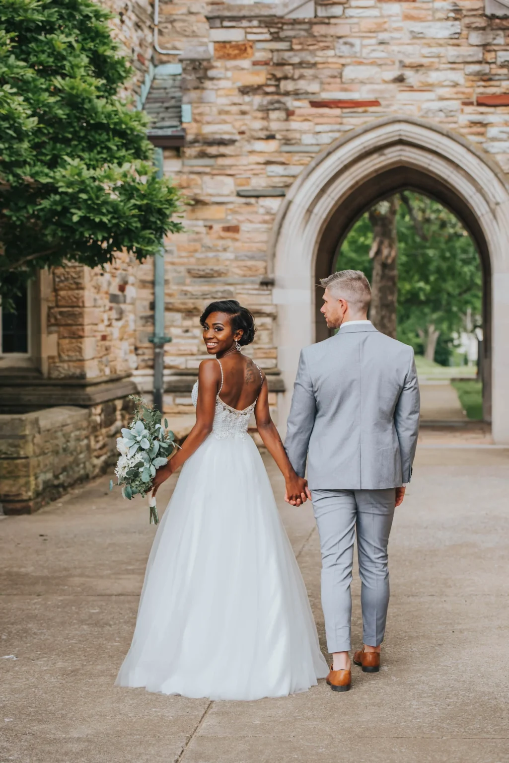 Elopement Wedding A bride and groom walk hand in hand away from the camera. The bride, wearing a white gown and holding a bouquet, looks back and smiles. The groom, dressed in a light gray suit, faces forward. They are outdoors in front of a stone archway and surrounded by greenery. Elopements Inc