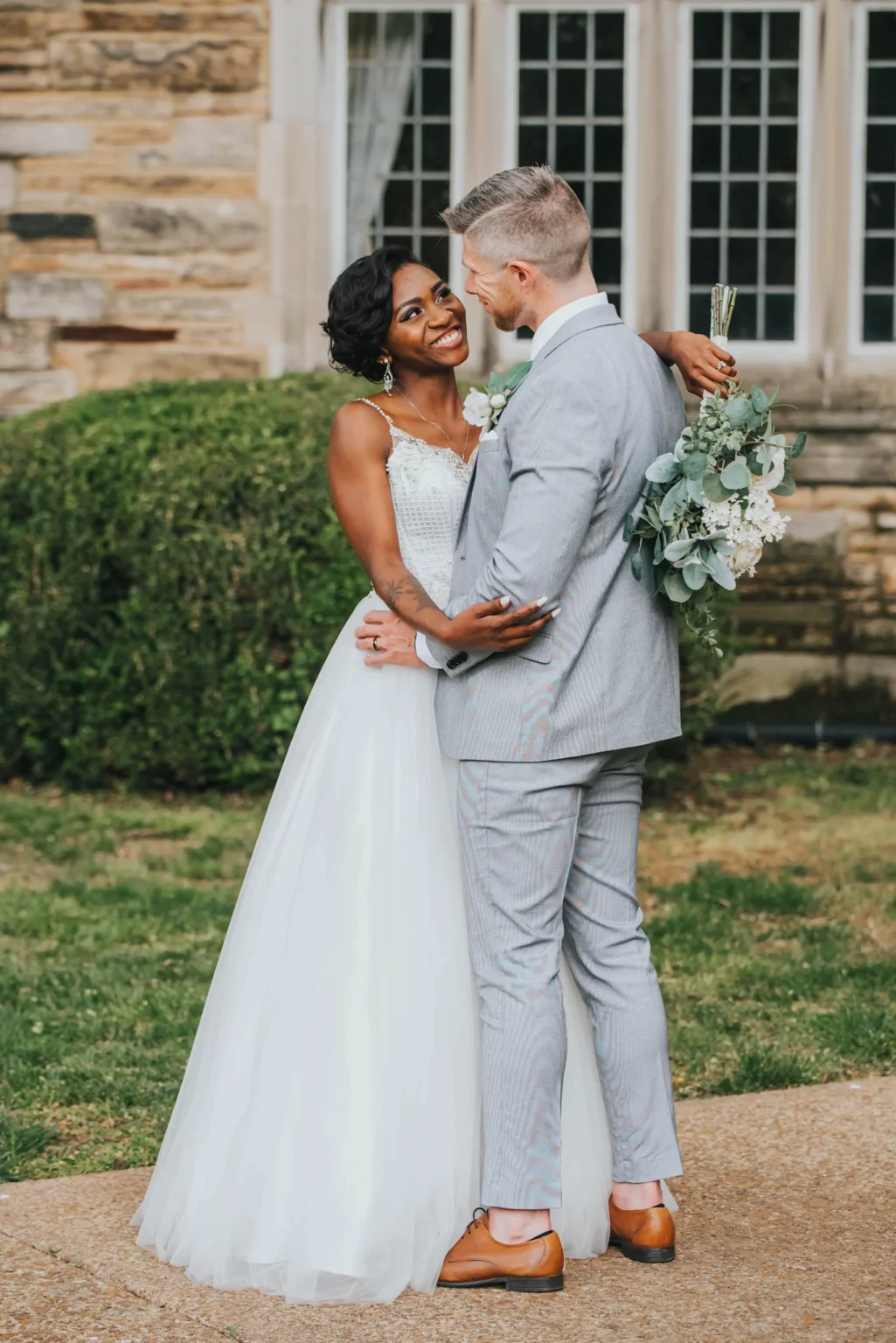 Elopement Wedding A newlywed couple stands intimately embracing outside a stone building with large windows. The bride, in an elegant white gown, smiles at the groom who wears a light gray suit. He's holding a bouquet of green and white flowers, and they both look happy and in love on their wedding day. Elopements Inc