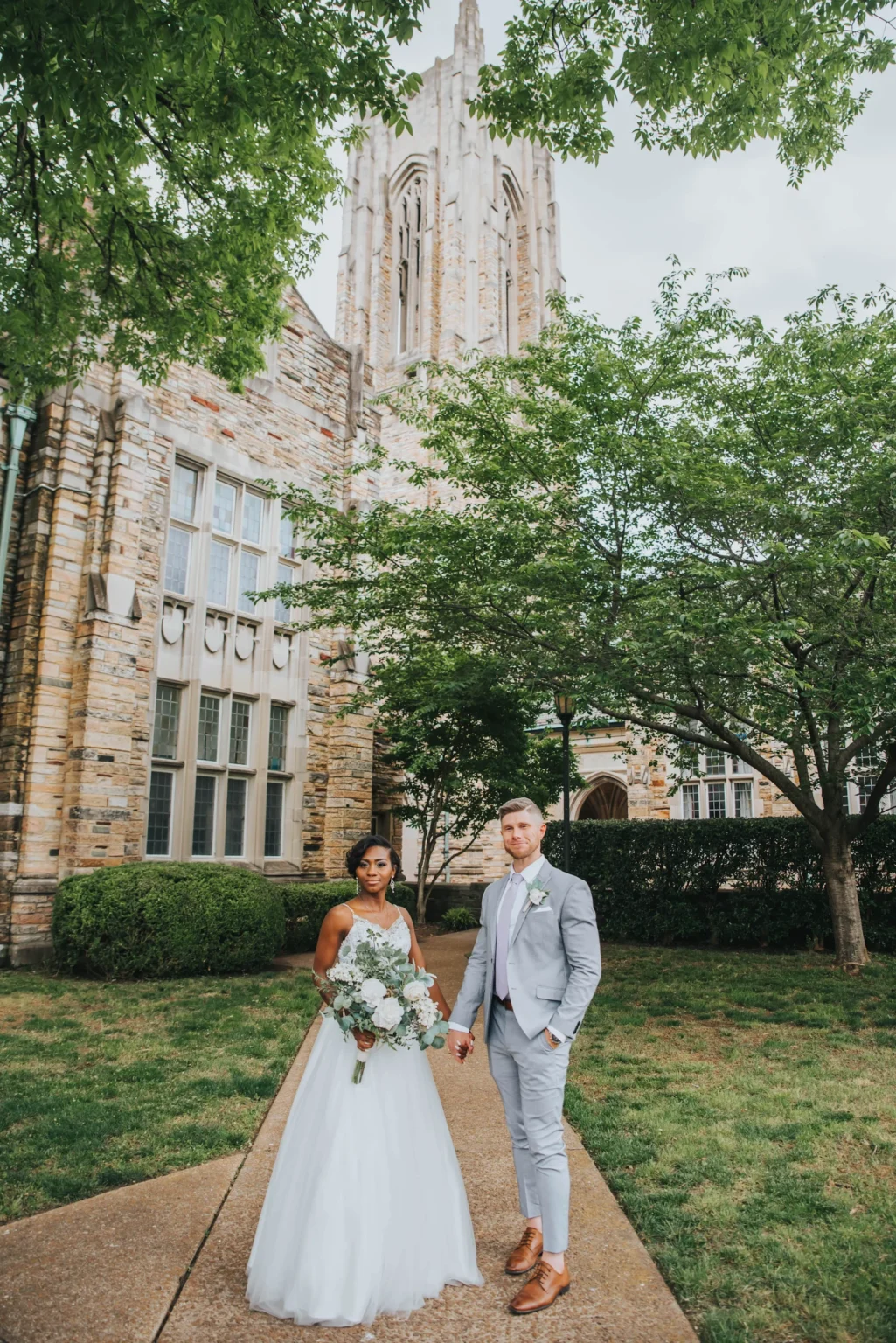 Elopement Wedding A newly-married couple stands hand in hand on a paved walkway in front of a historic stone building. The bride wears a white wedding gown and holds a bouquet of white flowers, while the groom is dressed in a light gray suit with a matching boutonniere. Green trees surround the scene. Elopements Inc