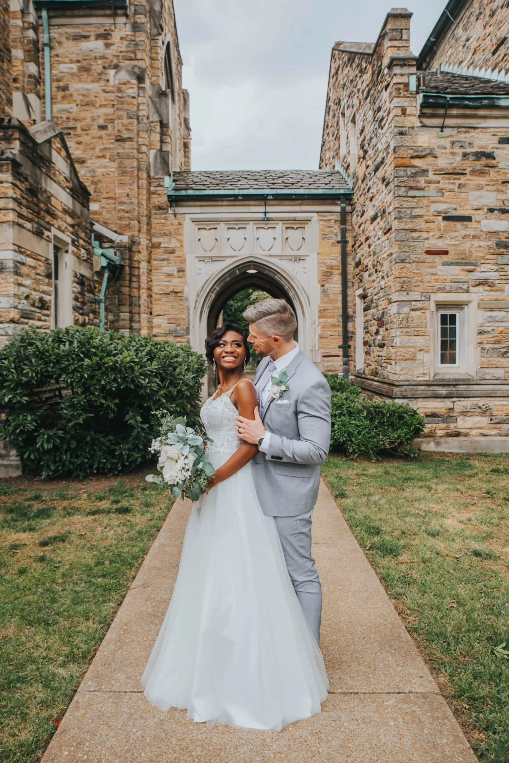 Elopement Wedding A bride and groom stand outside with greenery and a stone building in the background. The bride, in a white gown, holds a bouquet of flowers and smiles while looking up. The groom, in a light gray suit, stands behind her and looks at her lovingly, with his left arm around her waist. Elopements Inc