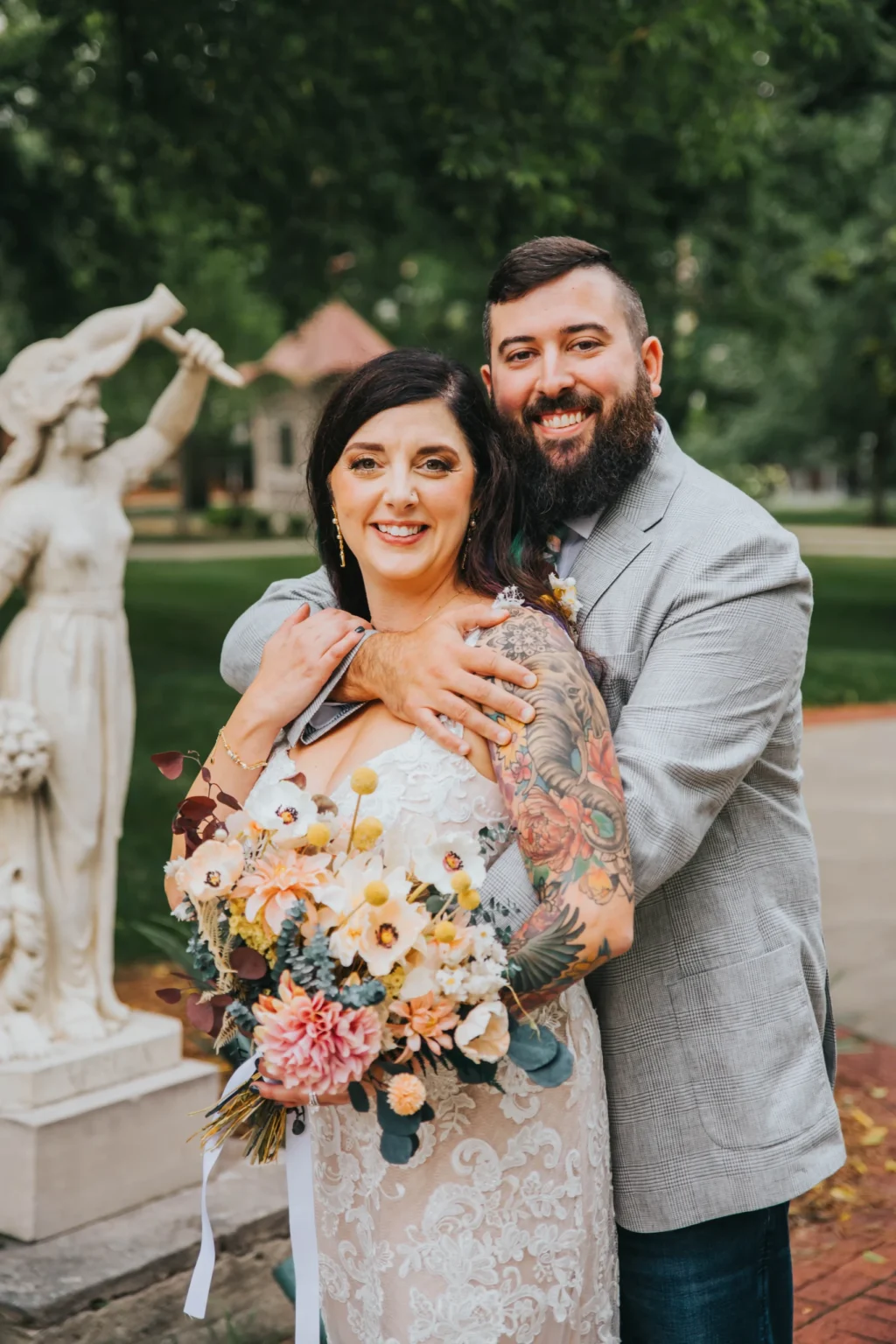 Elopement Wedding A couple poses outdoors in front of a statue. The woman, wearing a white lace dress and holding a bouquet of flowers, stands in front of the man who has his arms wrapped around her. The man is dressed in a light gray blazer and has a beard. Both are smiling and the background is lush and green. Elopements Inc