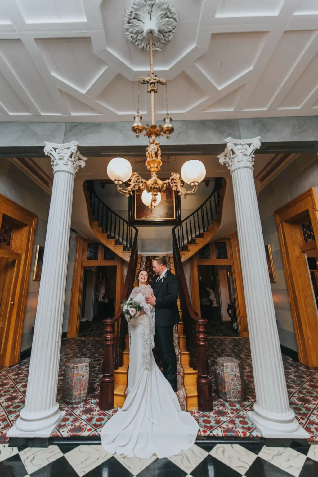 Elopement Wedding A newlywed couple stands together on a staircase in an elegant, ornately decorated room. The bride, in a white gown, holds a bouquet of flowers, and the groom, in a black suit, stands beside her. The grand staircase, white columns, and intricate ceiling light fixture create a sophisticated ambiance. Elopements Inc