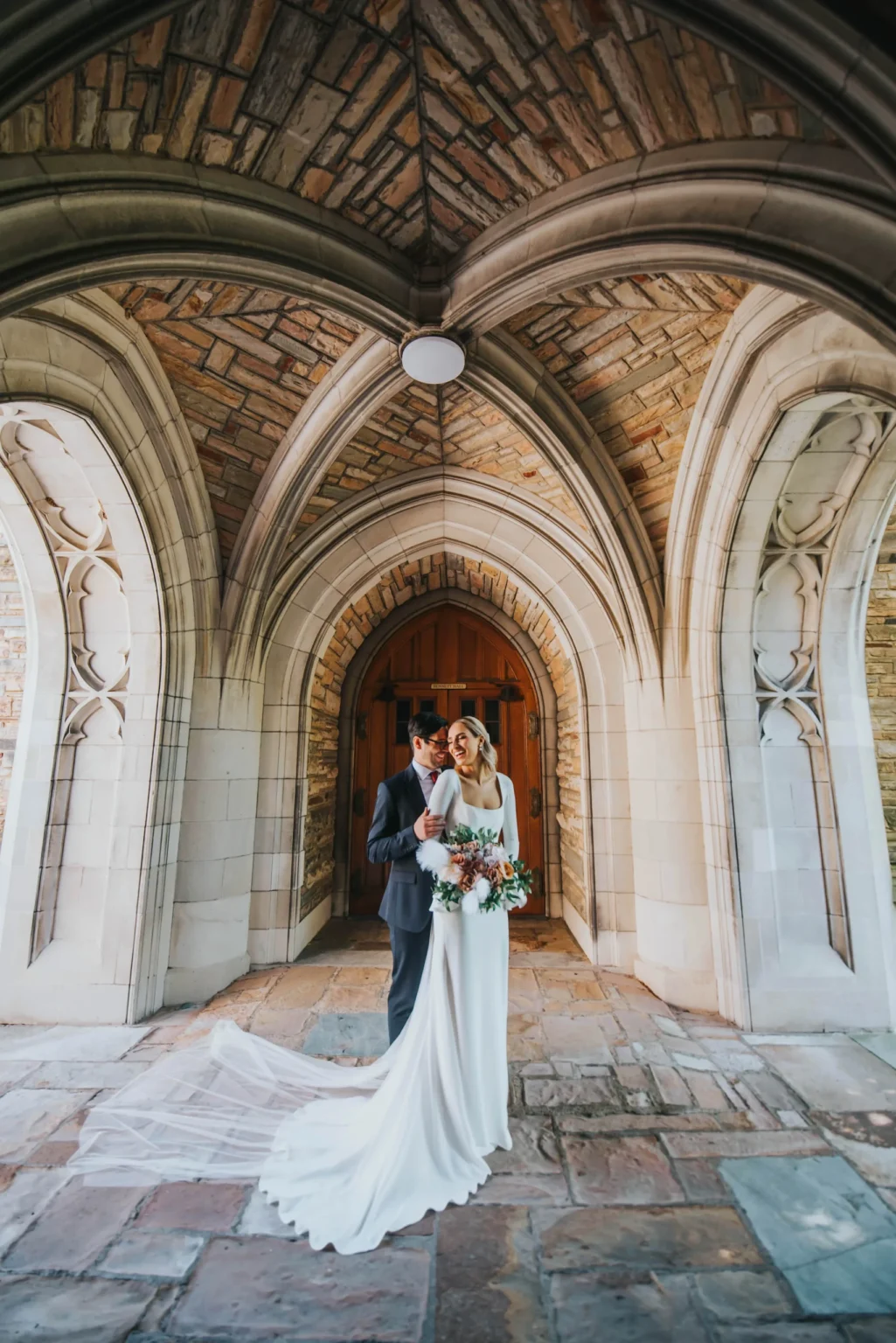 Elopement Wedding A bride and groom stand closely together under a stone archway with intricate vaulted ceilings. The bride is wearing a long white dress with a flowing train and holds a bouquet of white and pink flowers. The groom is dressed in a dark suit. They are standing on a stone pathway in front of wooden doors. Elopements Inc