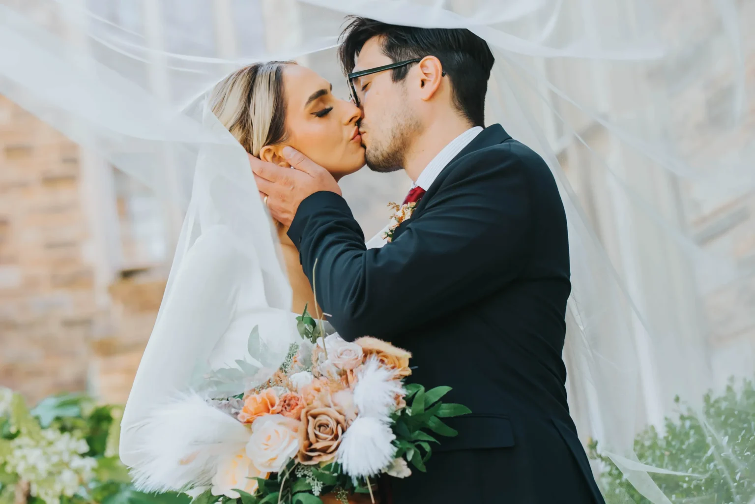 Elopement Wedding A bride and groom share a kiss on their wedding day. The bride, in a white wedding dress and veil, holds a bouquet of beige and pink flowers mixed with greenery. The groom, dressed in a black suit with glasses, gently touches her face. They stand in an outdoor setting with greenery. Elopements Inc