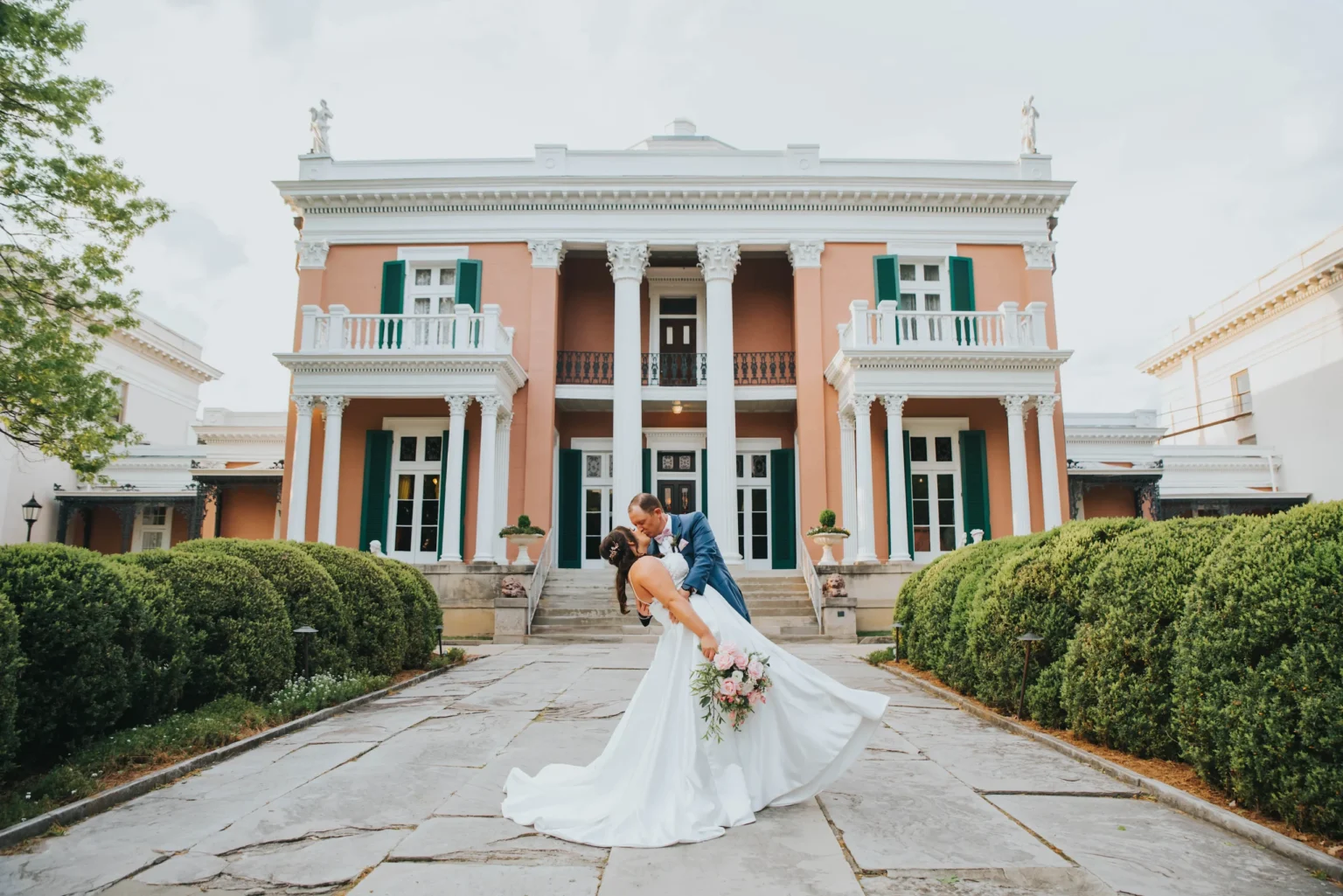 Elopement Wedding A groom in a blue suit dips and kisses a bride in a flowing white dress holding a bouquet, in front of an elegant, columned mansion with a peach facade and green shutters. Hedged bushes line the stone pathway leading up to the grand entrance of the house. Elopements Inc