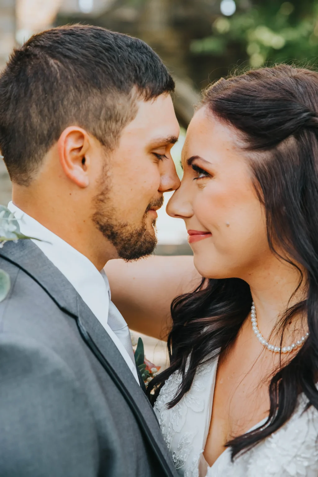 Elopement Wedding A couple stands closely together, face-to-face, smiling softly at each other. The man, wearing a gray suit and white tie, has short dark hair and a trimmed beard. The woman, with long brown hair, is adorned in a white dress and a pearl necklace, her hand resting gently on the man's shoulder. Elopements Inc