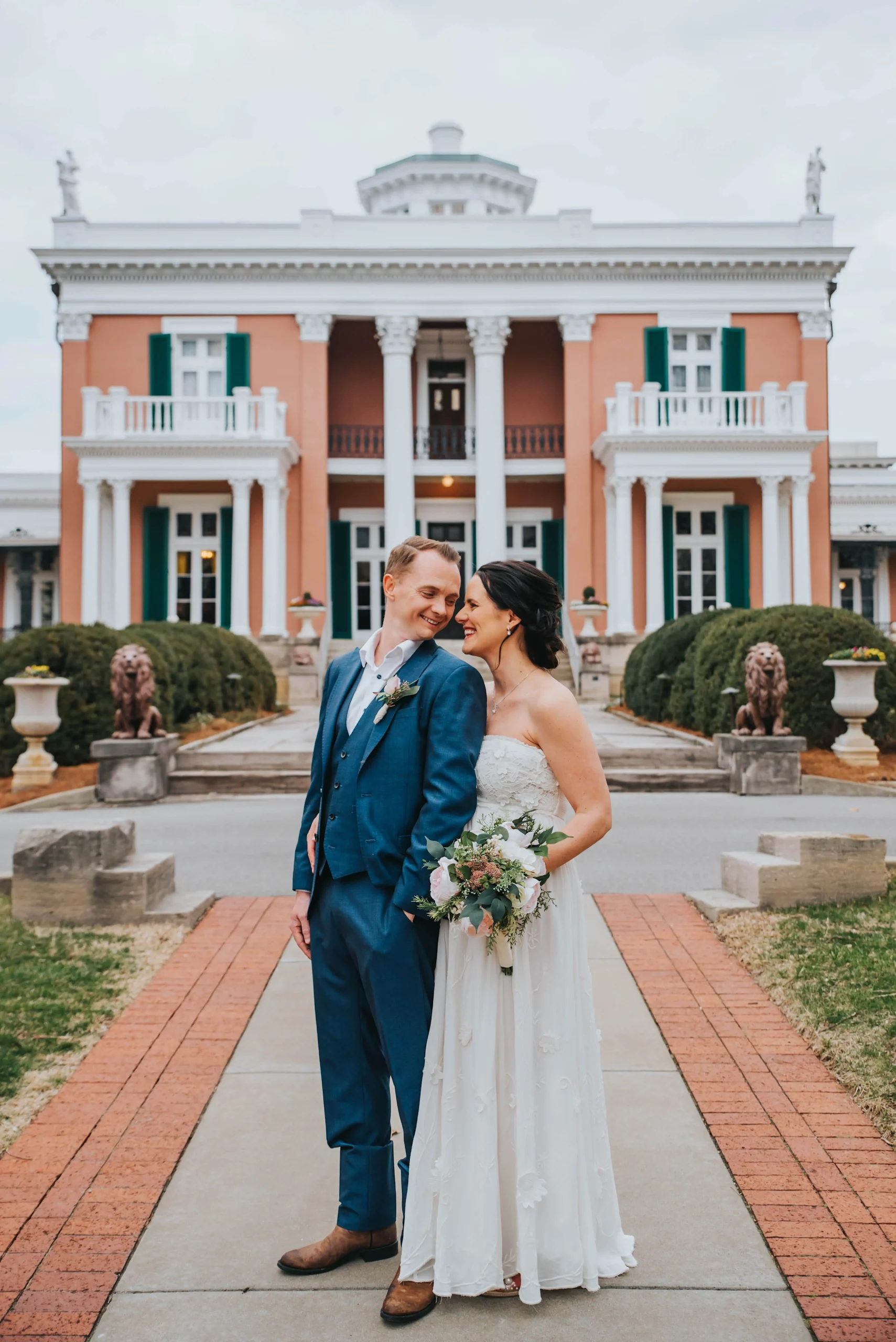 Elopement Wedding A smiling bride and groom pose in front of a grand building with white columns and green shutters. The groom, in a blue suit and brown shoes, leans slightly towards the bride, who is in a white strapless gown, holding a bouquet of flowers. The path is flanked by stone lion statues and greenery. Elopements Inc