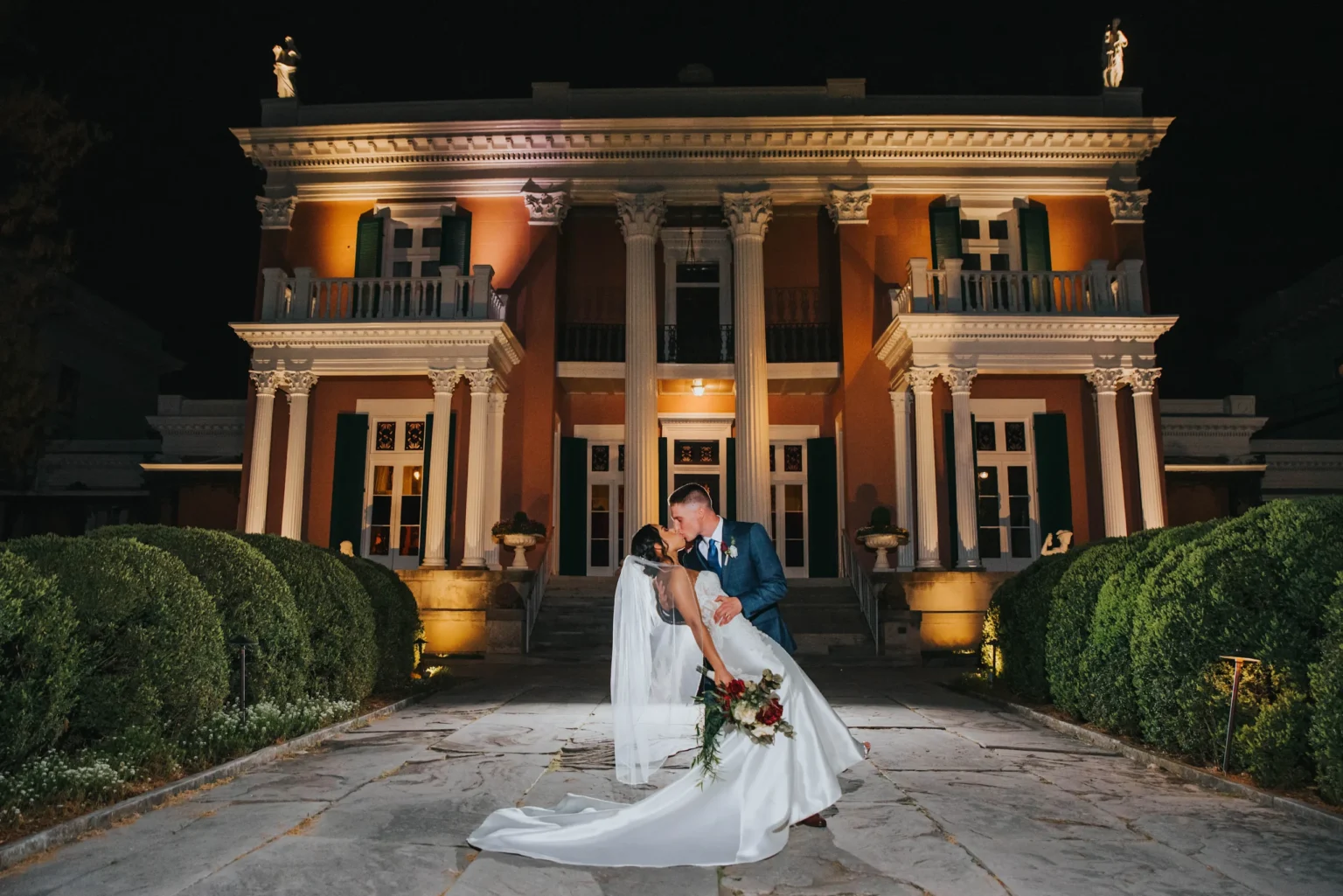 Elopement Wedding A bride and groom share a romantic kiss in front of a grand, illuminated building with columns and statues at night. The bride holds a bouquet of red and white flowers and wears a long, flowing white gown with a veil. The groom is in a blue suit, leaning her back gently as they kiss. Elopements Inc