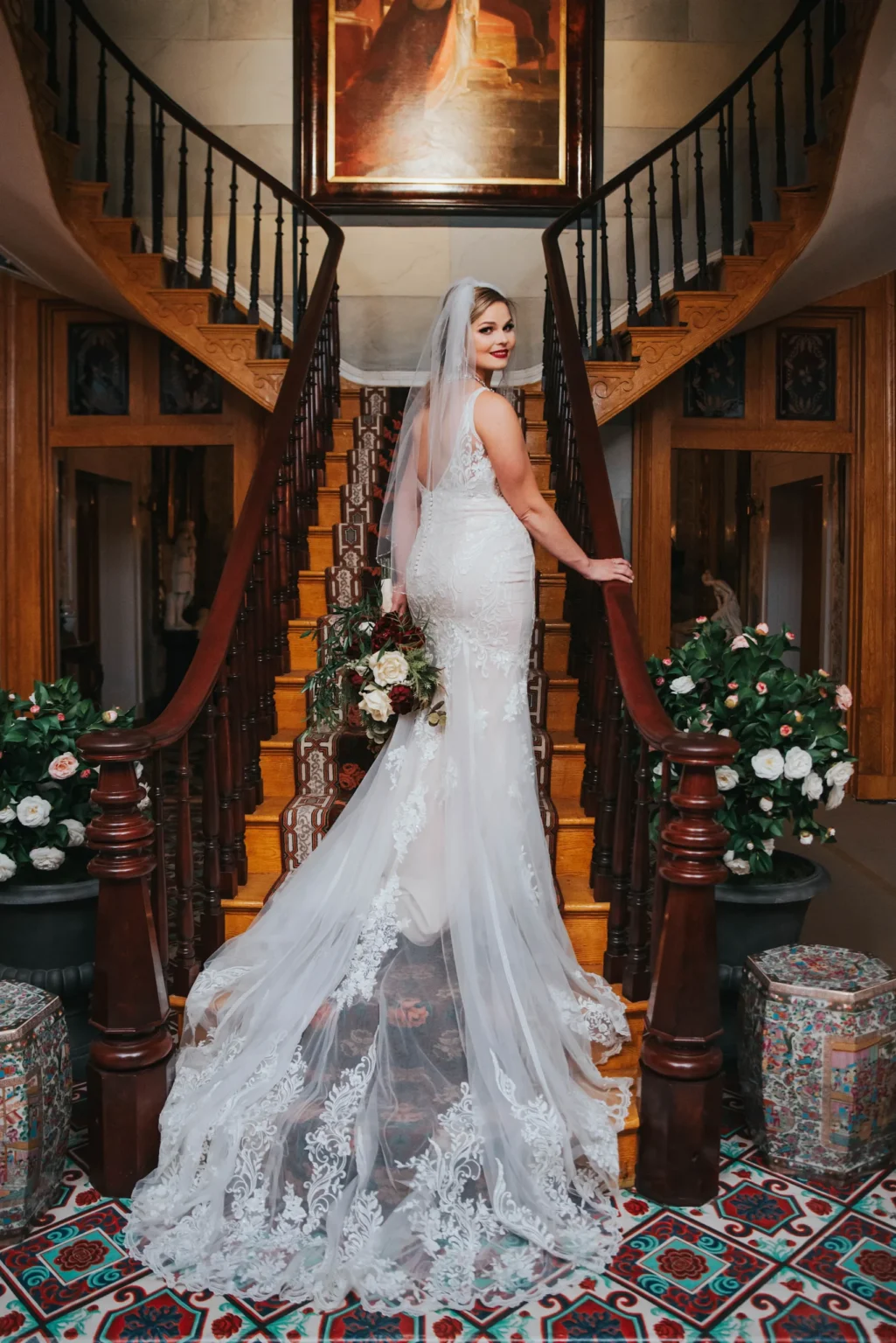 Elopement Wedding A bride in a white, lace wedding gown and veil stands on the first step of an ornate, wooden spiral staircase, looking back over her shoulder. She holds a bouquet of flowers. The staircase is flanked by lush flower arrangements and decorative rugs. A large framed painting hangs above the landing. Elopements Inc