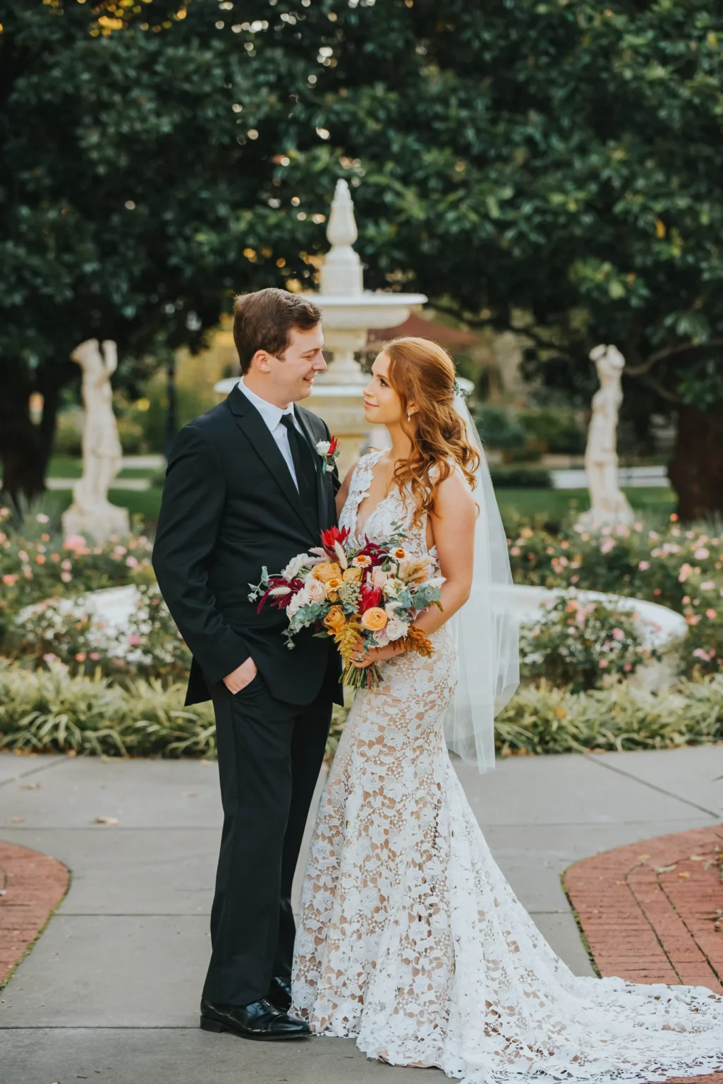 Elopement Wedding A bride and groom stand facing each other outdoors on a paved path. The bride, in a lacy white wedding dress, holds a bouquet of colorful flowers and has a long veil trailing behind her. The groom wears a black suit with a white shirt and black tie. Behind them is a lush garden with a white statue. Elopements Inc