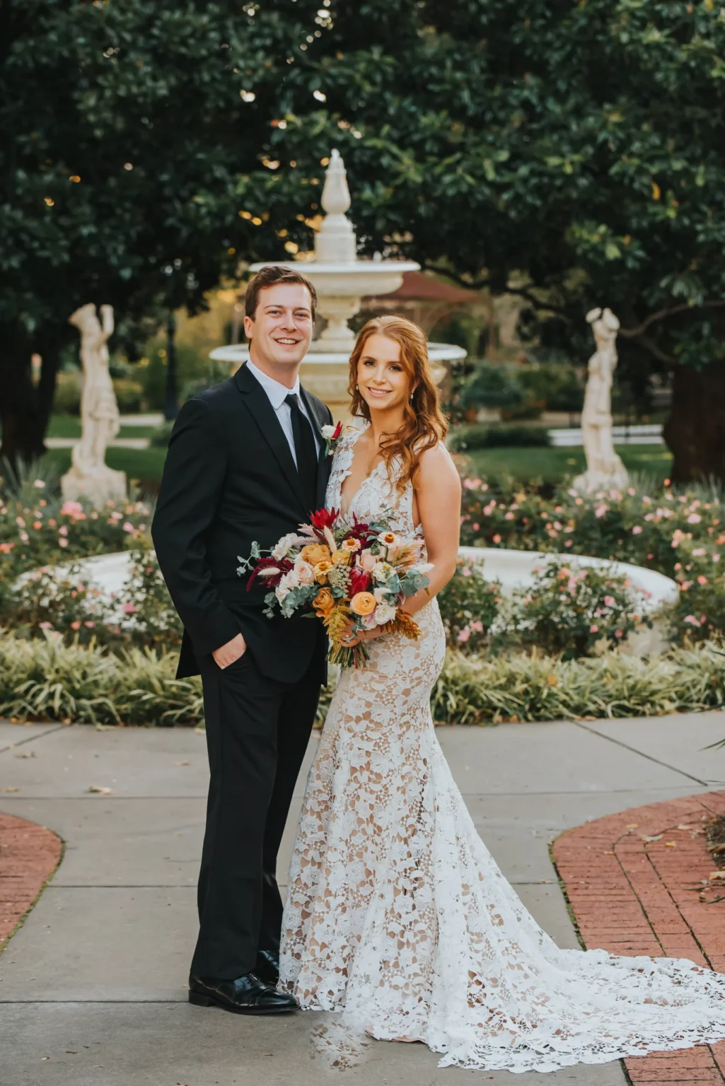 Elopement Wedding A couple stands together in an outdoor garden setting. The man wears a black suit with a white shirt and black tie. The woman wears a lace wedding dress, holding a bouquet of colorful flowers. They both look at the camera and smile. A fountain and statues are visible in the background. Elopements Inc