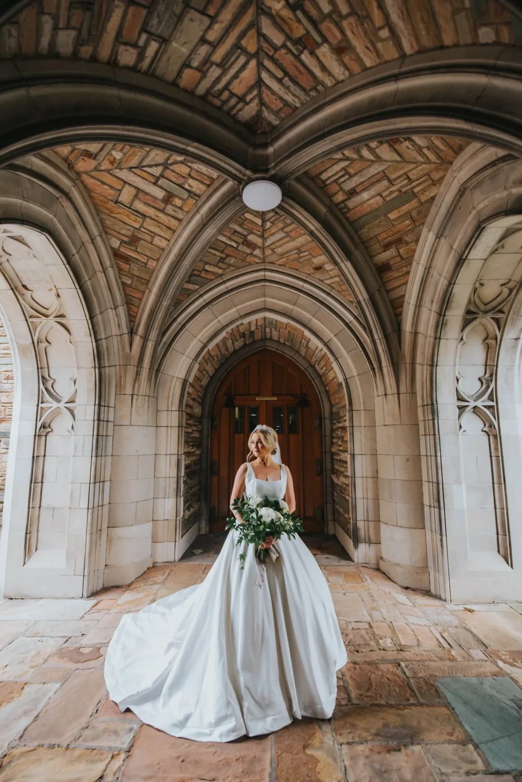 Elopement Wedding A bride in a flowing white gown stands alone under a stone archway. She is holding a bouquet of green and white flowers. The archway features intricate stonework and a wooden door in the background. The floor beneath her is made of large stone tiles. Elopements Inc