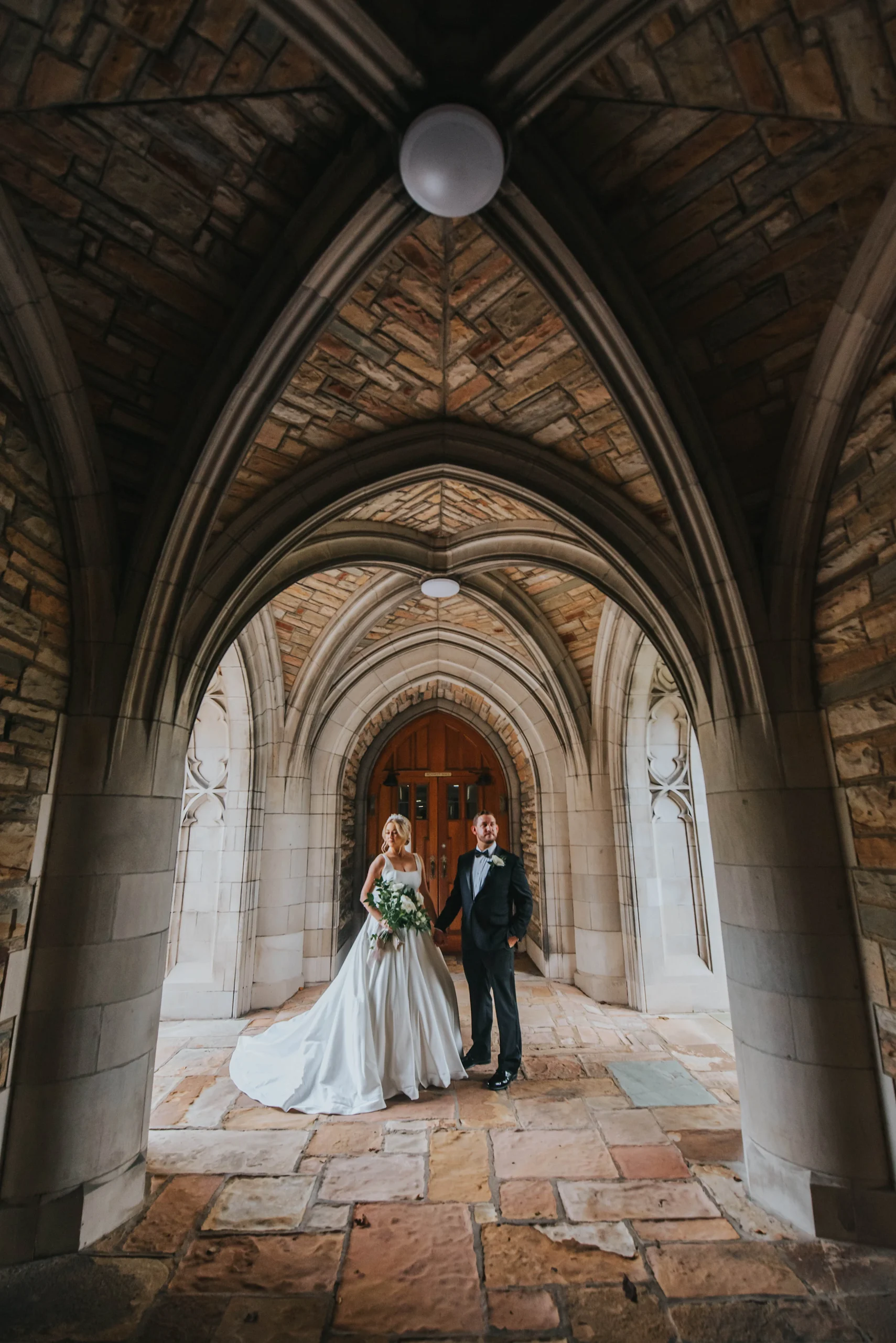 Elopement Wedding A bride in a flowing white gown and a groom in a black suit stand under a stone archway with Gothic architecture. The arched ceiling above them features intricate stonework. They pose on a stone pathway, illuminated by soft lighting, near a wooden door framed by more stone arches. Elopements Inc