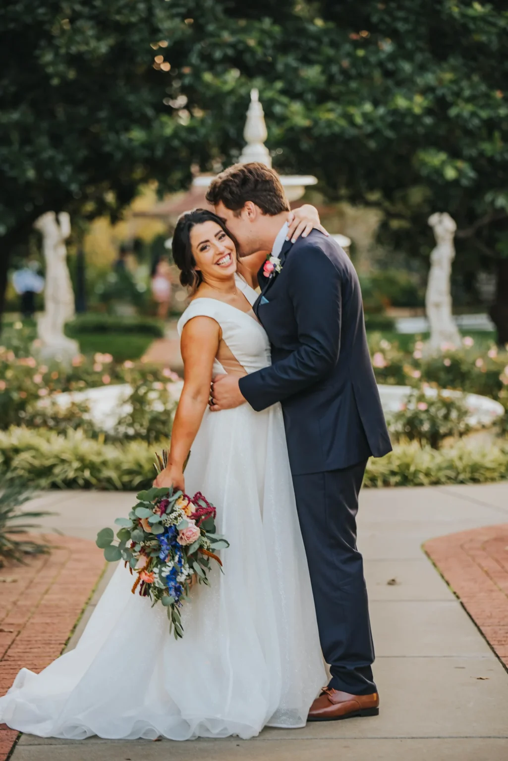 Elopement Wedding A bride in a white dress holds a colorful bouquet and leans back, smiling as her groom embraces her from the side. The groom, in a navy suit, has his arms around her and kisses her cheek. They stand on a walkway in a lush garden with greenery and statues in the background. Elopements Inc