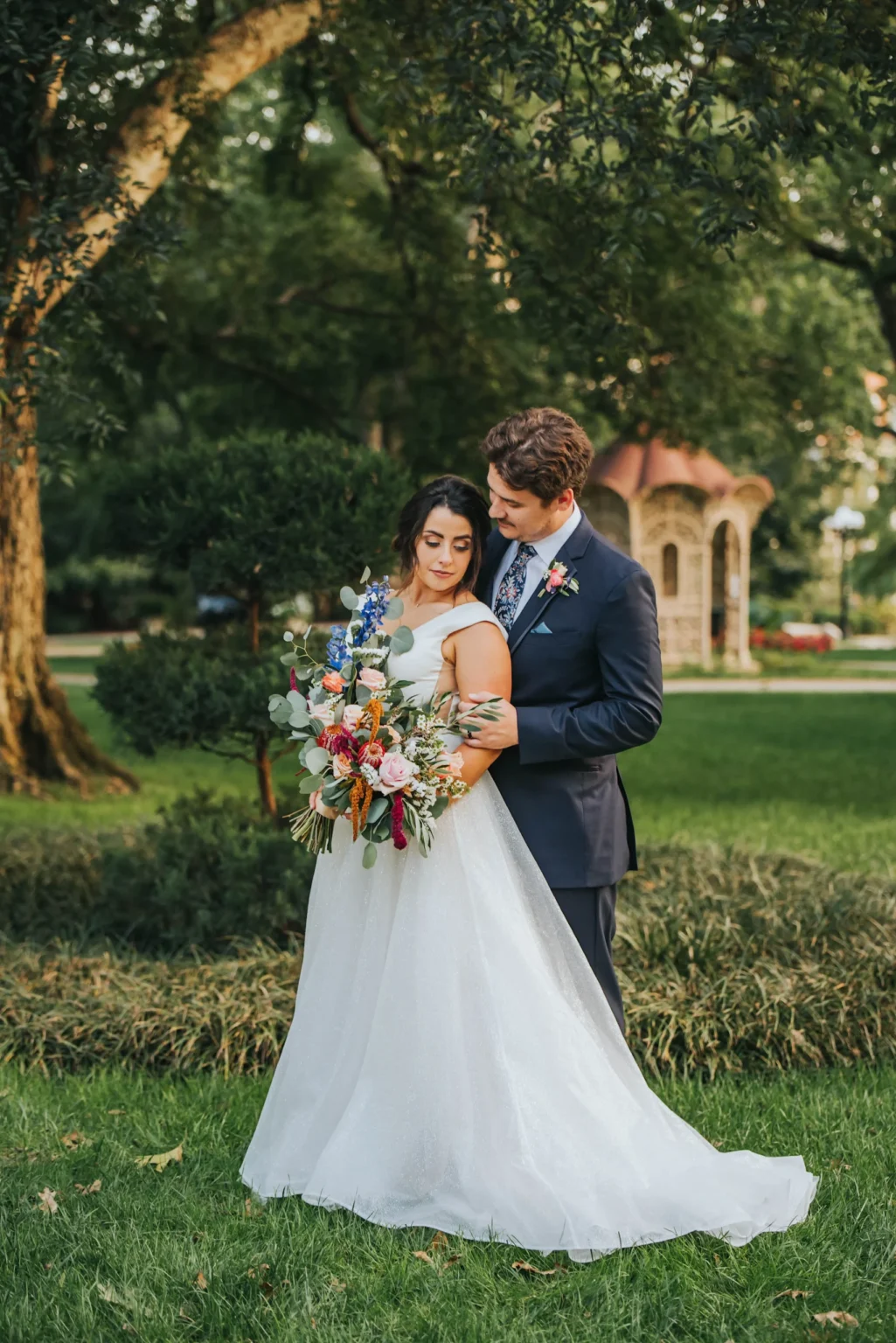 Elopement Wedding A bride in a white gown holds a colorful bouquet while leaning against a groom dressed in a navy suit. They stand in a verdant park with lush grass, trees, and a quaint gazebo in the background. The couple shares an intimate moment, with the groom gently holding the bride from behind. Elopements Inc