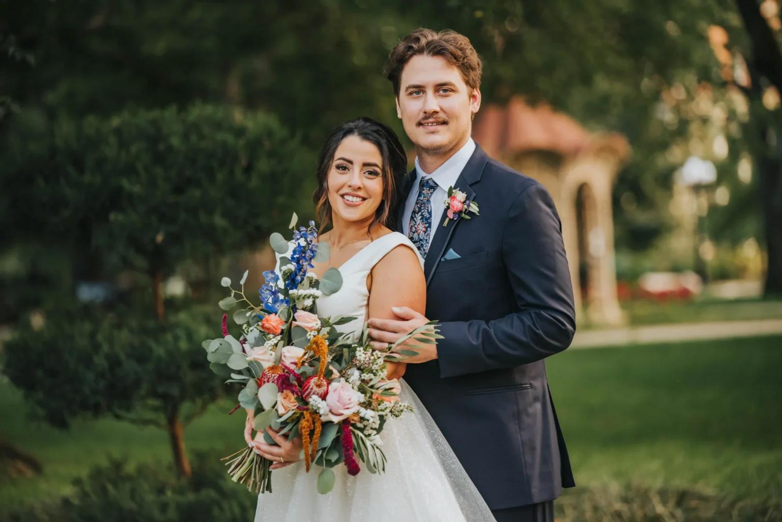 Elopement Wedding A bride and groom are outdoors, standing closely together. The bride is holding a vibrant bouquet with various flowers, including blue, pink, and orange. She is wearing a white gown, and the groom is in a navy suit with a floral tie and boutonnière. There is greenery and an archway in the background. Elopements Inc