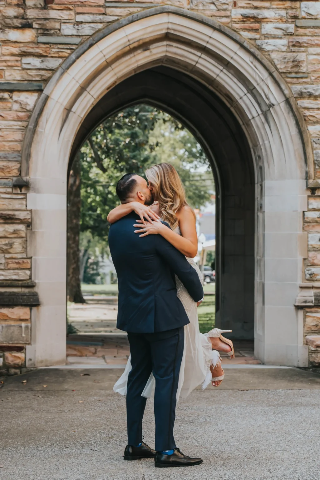 Elopement Wedding A couple dressed in formal attire embrace under a stone archway. The groom in a blue suit lifts the bride in a white dress as they share a kiss. The scene is set outdoors with greenery visible in the background through the arch. The bride's heels dangle as they hold each other closely. Elopements Inc