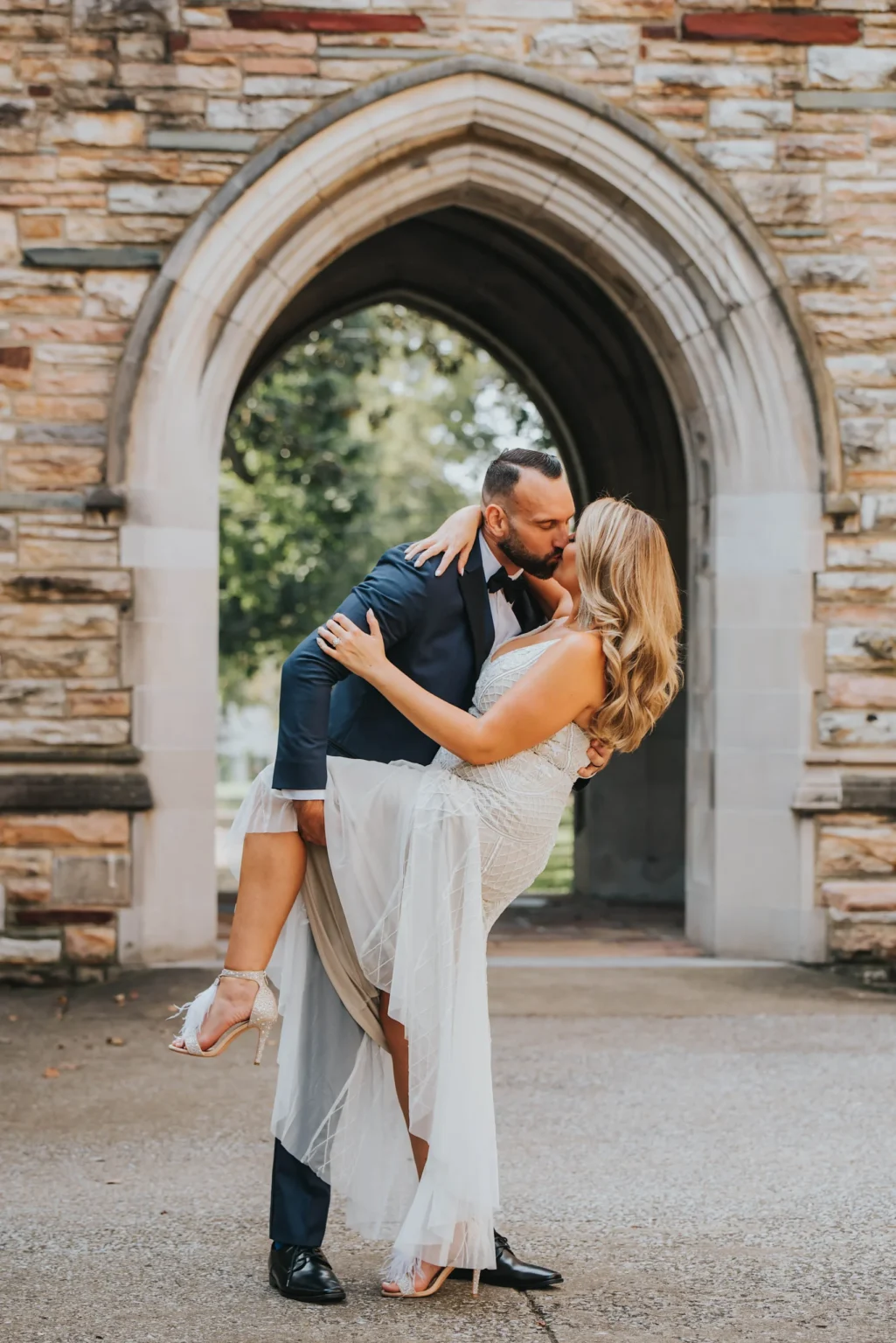 Elopement Wedding A man dressed in a dark blue suit and a woman in a white dress share a romantic kiss. The man holds the woman in a dip, her leg bent and lifted, against a backdrop of a stone archway with greenery visible through the arch. The scene exudes love and elegance. Elopements Inc