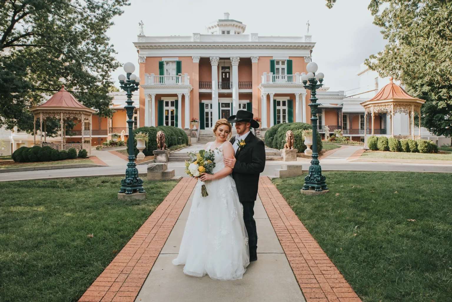 Elopement Wedding A bride and groom stand on a paved pathway in front of a large, elegant mansion. The bride is in a white gown holding a bouquet, and the groom is wearing a black suit and hat. The red brick mansion features white columns, green shutters, and ornate details. Lush greenery surrounds the scene. Elopements Inc