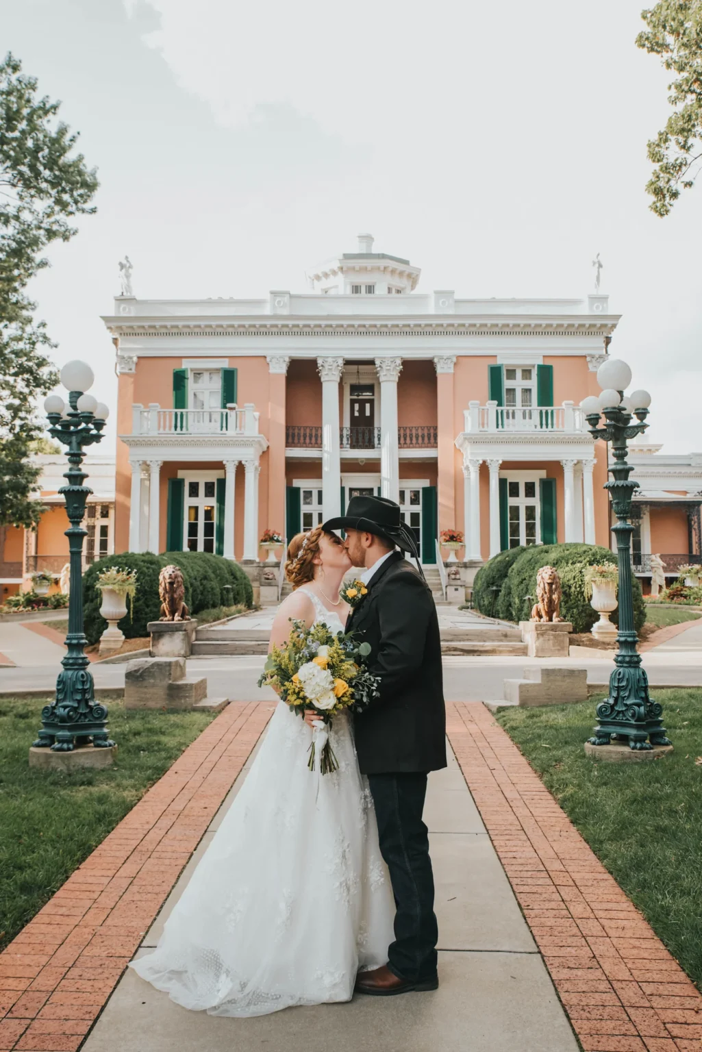 Elopement Wedding A bride and groom share a kiss in front of a grand, historic mansion adorned with white columns and green shutters. The bride wears a white gown and holds a bouquet of yellow flowers. The groom wears a black cowboy hat and suit. They stand on a red-brick path flanked by ornate lampposts and manicured shrubs. Elopements Inc