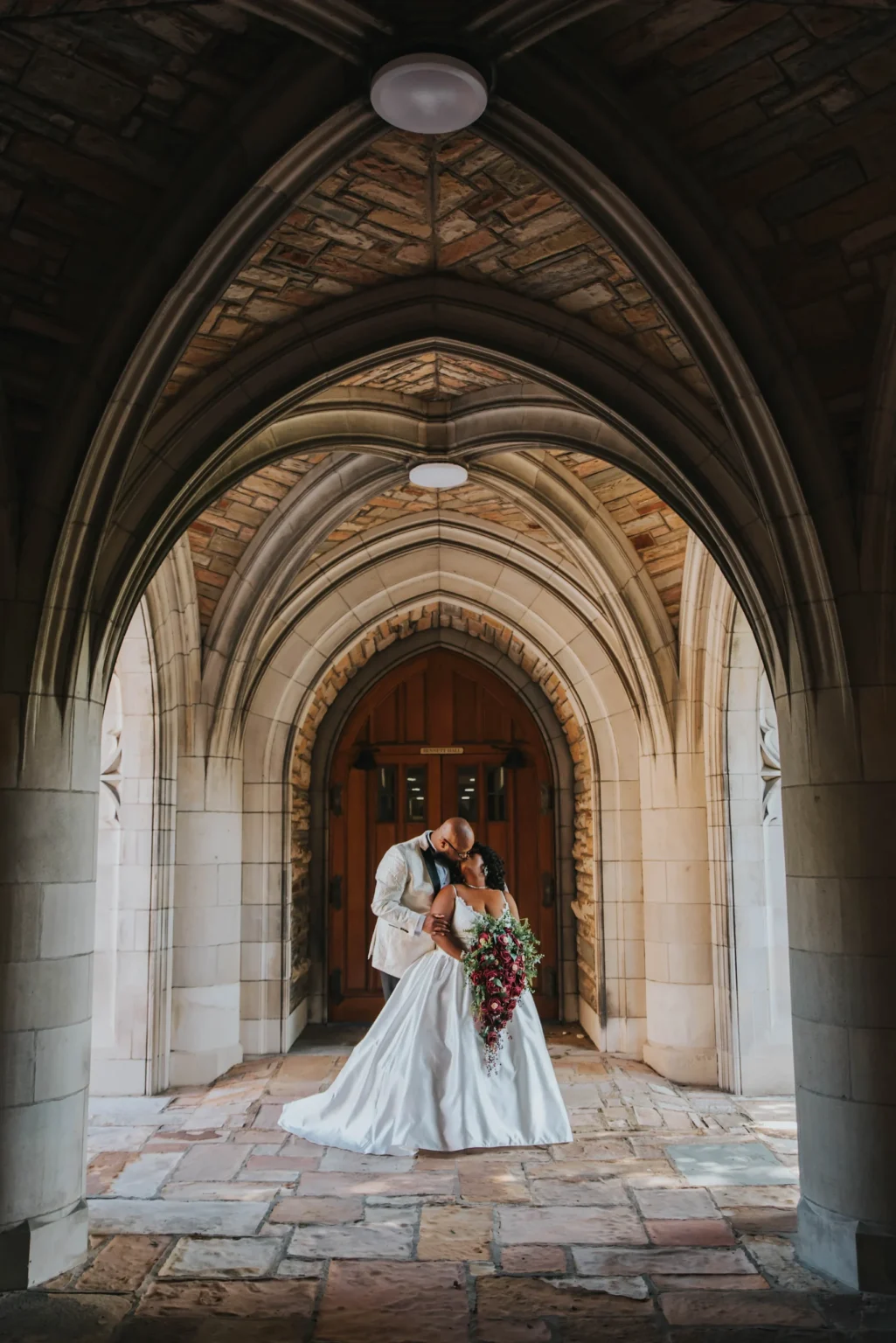 Elopement Wedding A newlywed couple stands in an embrace under a series of stone archways at a gothic-style venue. The bride, in a flowing white gown, holds a vibrant, cascading bouquet. The groom, in a white jacket and black pants, lovingly kisses her forehead. Light filters through the arches, adding a warm glow. Elopements Inc