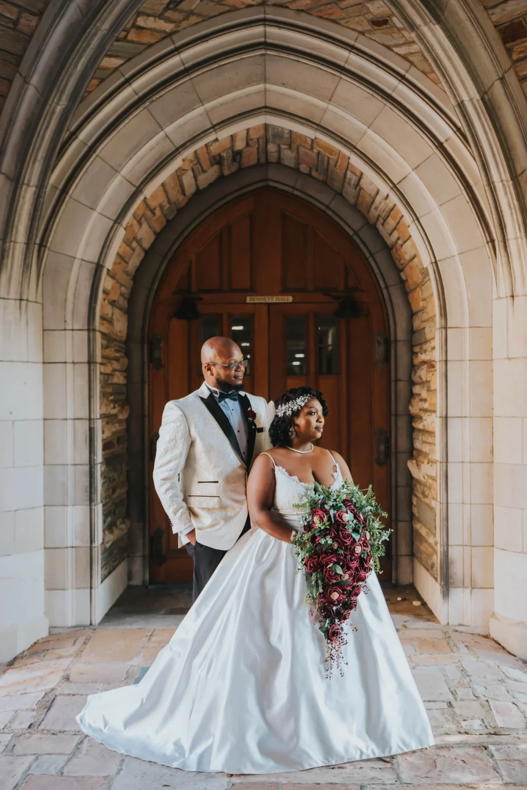 Elopement Wedding A newlywed couple stands under a stone archway in front of a wooden door. The groom wears a white tuxedo jacket and black bow tie, while the bride wears a white, flowing gown and holds a cascading bouquet of deep red and green flowers. The stonework and architectural details frame the couple elegantly. Elopements Inc