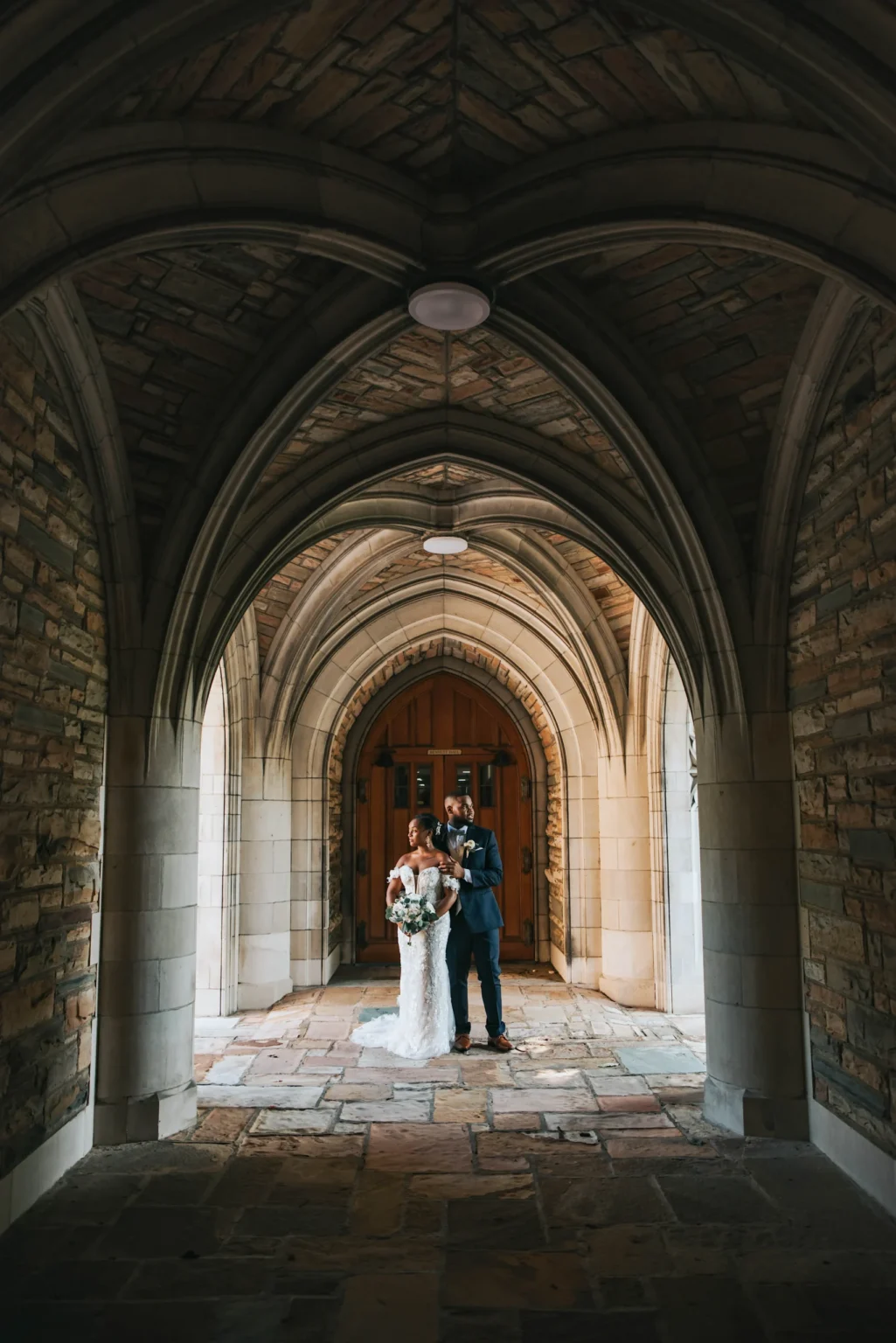 Elopement Wedding A bride in a white dress with a bouquet and a groom in a dark suit stand under a stone vaulted archway. Natural light illuminates them from behind, casting soft shadows on the stone walls and floor. The background features a closed wooden door framed by the architectural elements. Elopements Inc