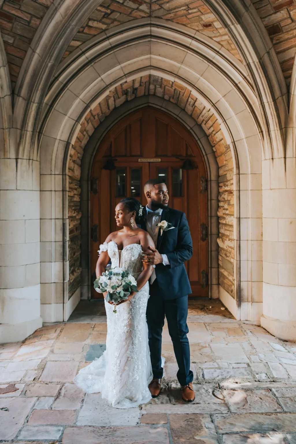 Elopement Wedding A bride and groom stand under a stone archway with wooden double doors behind them. The bride wears a white, off-the-shoulder lace gown and holds a bouquet of flowers. The groom wears a dark suit with a boutonnière and stands closely behind her, looking to the side. Both appear serene and composed. Elopements Inc