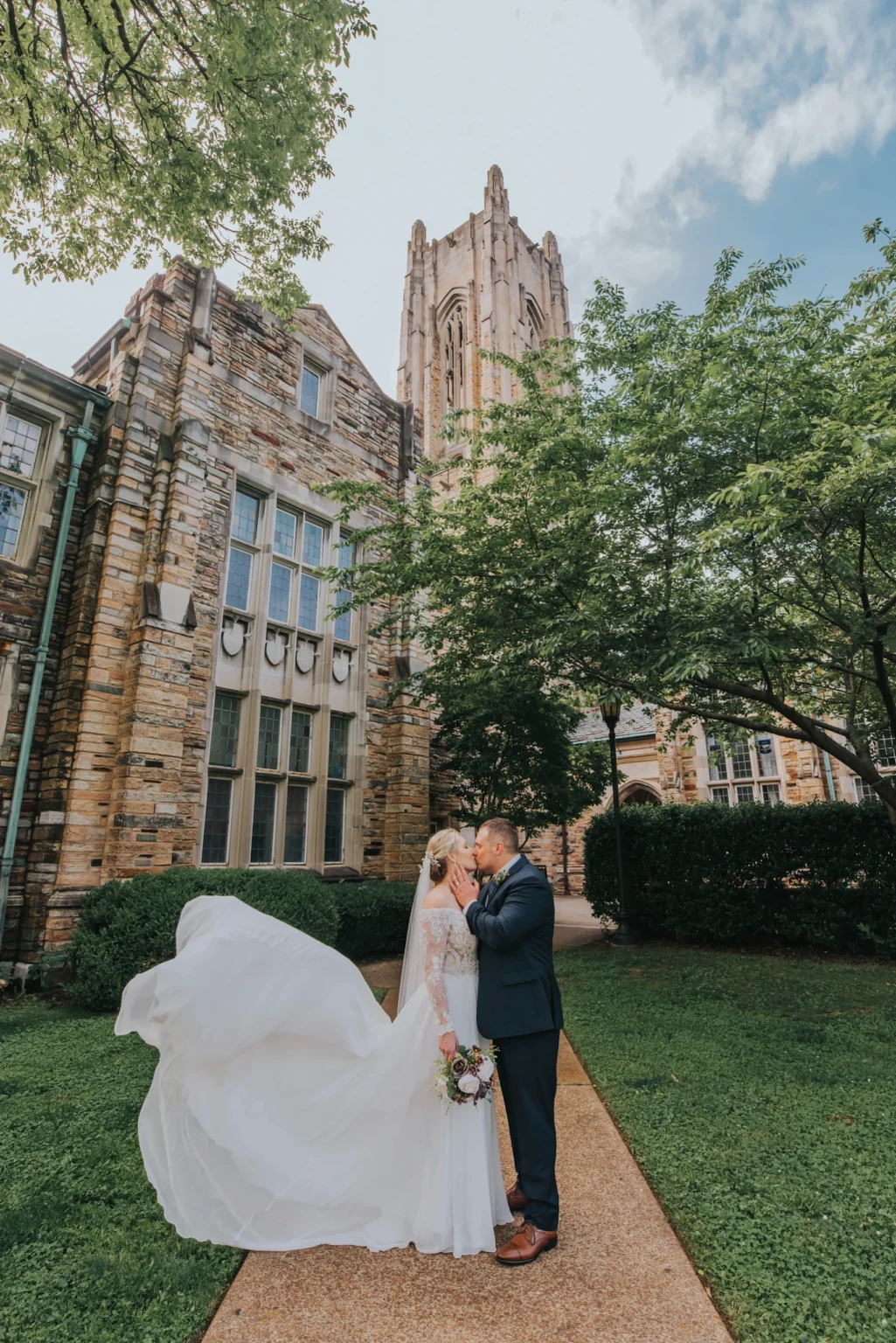 Elopement Wedding A bride and groom share a kiss outside a historic stone building with arched windows and a tall, pointed tower. The bride wears a flowing white gown, and the groom is in a dark suit. They are surrounded by lush green trees and grass on a sunny day. Elopements Inc