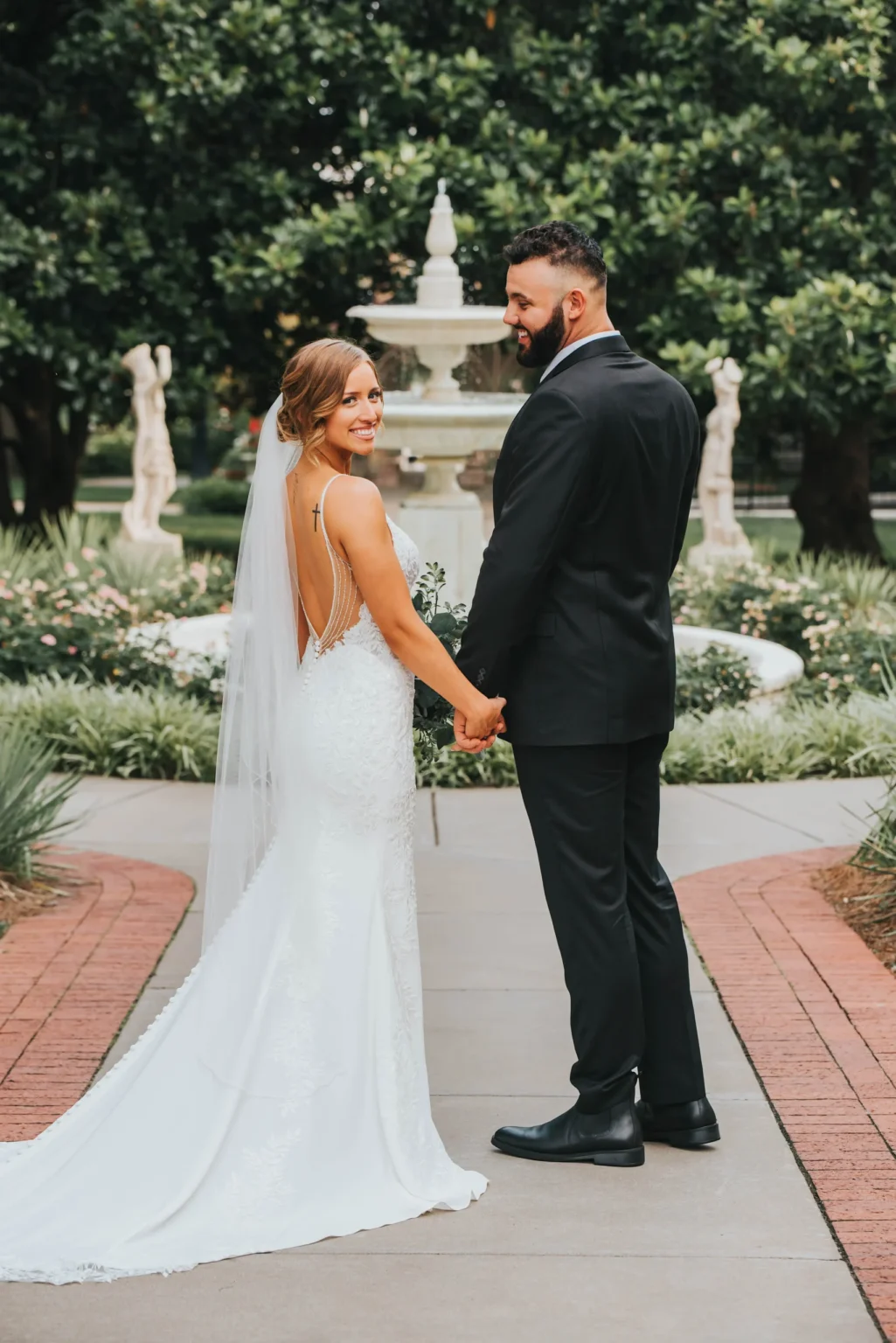 Elopement Wedding A bride and groom stand hand in hand on a paved path, facing toward a fountain surrounded by greenery. The bride wears a white, backless gown with a long train and veil, and the groom dons a black suit. They both smile, looking over their shoulders at the camera. Elopements Inc