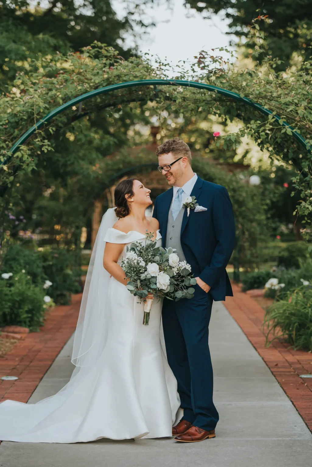 Elopement Wedding A bride in a white dress and veil stands closely beside a groom in a navy blue suit and tie, both smiling at each other. The bride holds a bouquet of white flowers and greenery. They stand under a garden arch adorned with greenery, with a brick pathway and lush greenery around them. Elopements Inc