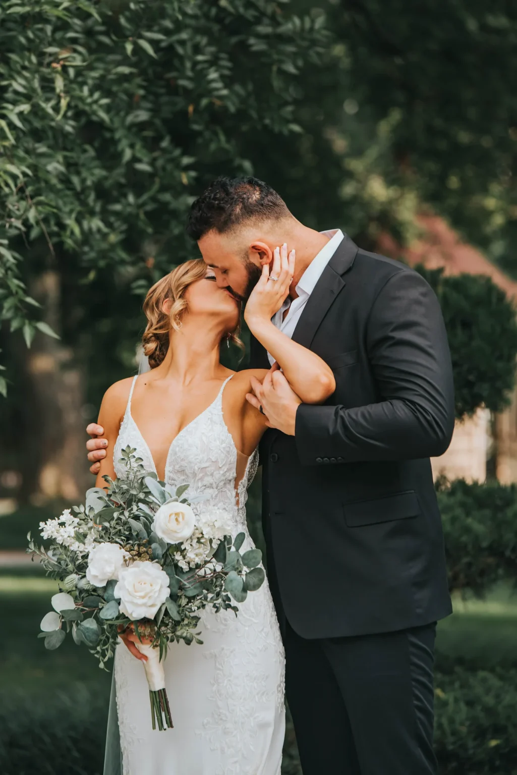 Elopement Wedding A bride and groom are kissing outdoors. The bride wears a white lace gown and holds a bouquet of white flowers and greenery. The groom wears a black suit and gently holds the bride's face with his hand. They are surrounded by lush green trees and foliage, creating a serene background. Elopements Inc