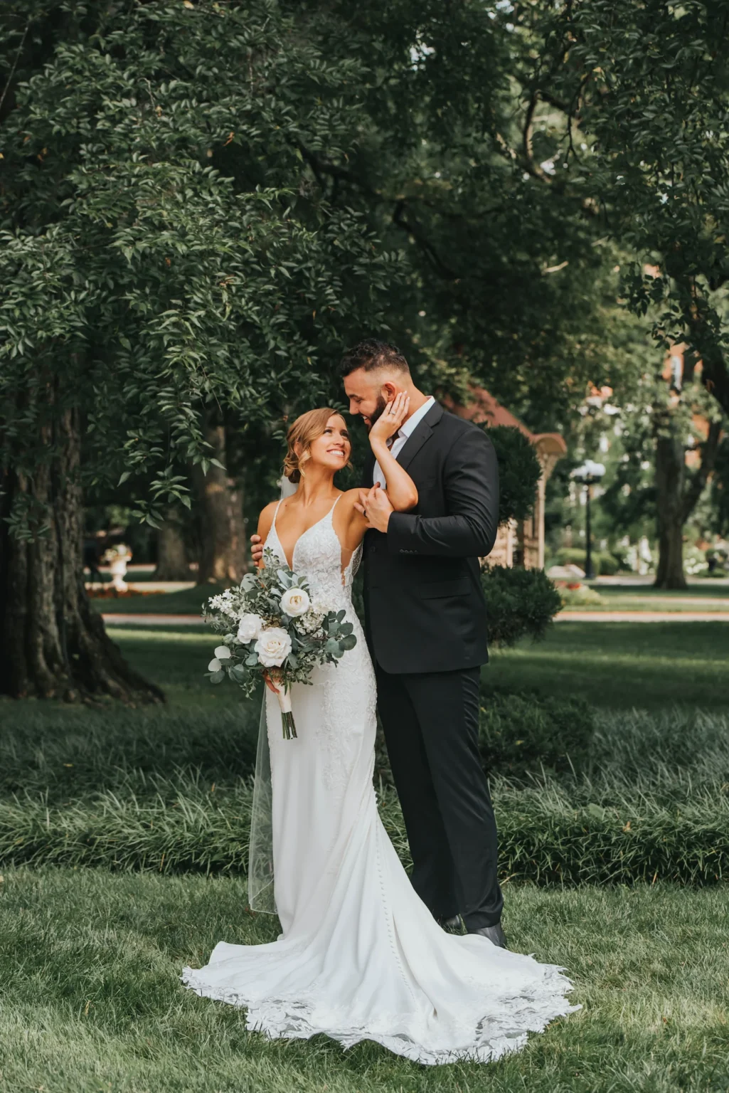 Elopement Wedding A bride and groom stand on a lush green lawn surrounded by tall trees. The bride is wearing a white dress with lace details and holding a bouquet of white and greenery flowers. The groom, in a black suit, gently touches her face as they smile at each other. They appear joyful and content. Elopements Inc