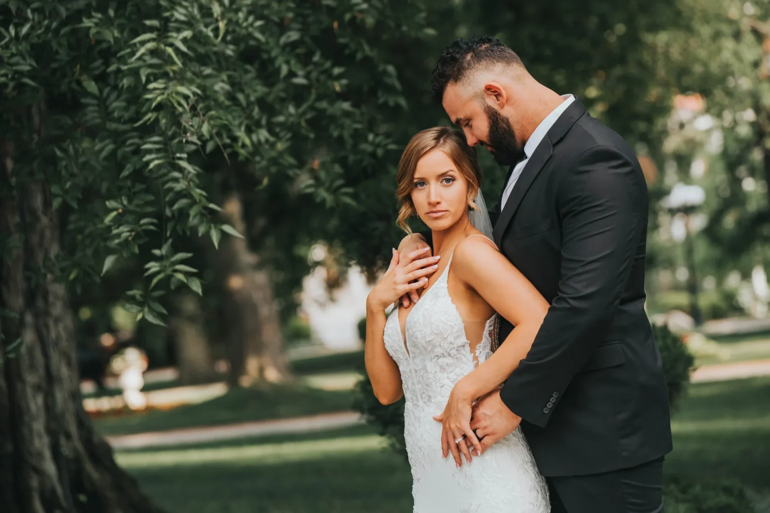 Elopement Wedding A couple stands closely in an outdoor setting. The woman, in a white lace wedding dress, looks towards the camera with her hand touching her chest, while the man, in a black suit, gently embraces her from behind. They are surrounded by lush green trees and soft sunlight filters through the leaves. Elopements Inc