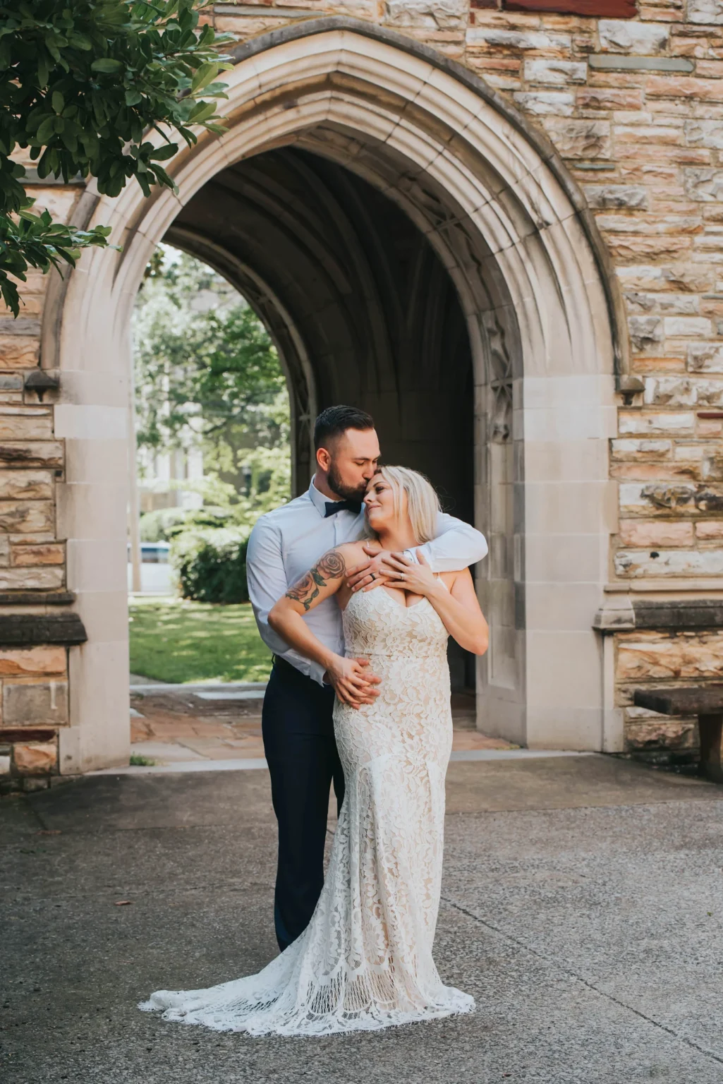 Elopement Wedding A couple stands in a loving embrace under a stone archway. The man, wearing a light blue shirt and dark pants, holds the woman from behind. She is in a sleeveless, lace-patterned white dress. They are both smiling, with a green, lush park visible through the archway behind them. Elopements Inc