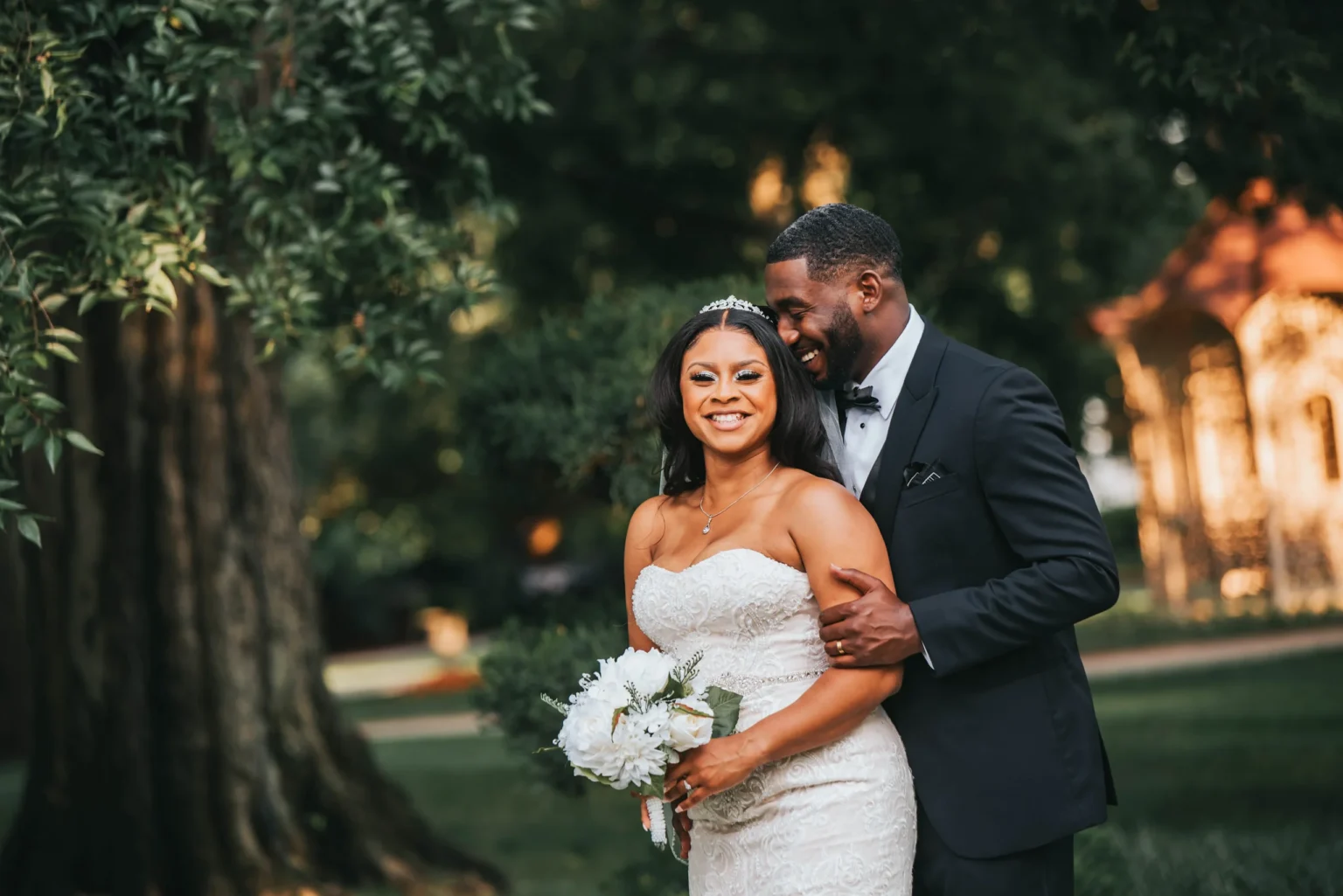 Elopement Wedding A joyful bride and groom stand outdoors surrounded by lush greenery. The bride, in a strapless white dress with a bouquet, smiles brightly. The groom, in a black tuxedo, stands behind her with a cheerful expression. Sunlight filters through trees in the background, creating a warm atmosphere. Elopements Inc