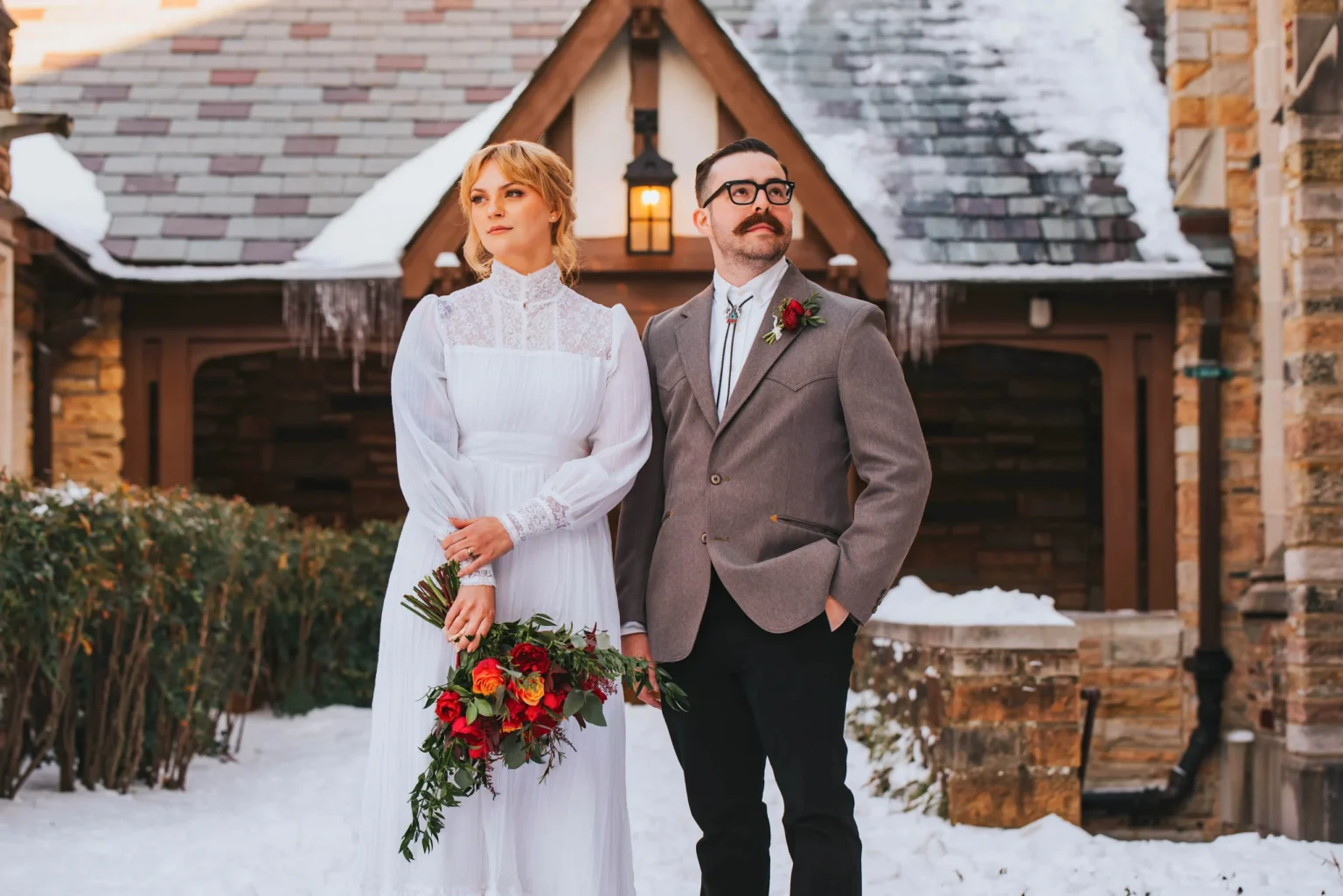 Elopement Wedding A couple stands in a snowy outdoor setting in front of a stone building with triangular roofs. The woman, wearing a white, long-sleeved lace dress, holds a colorful bouquet of red and orange flowers. The man, dressed in a gray jacket, white shirt, and black pants, stands with a confident posture. Elopements Inc