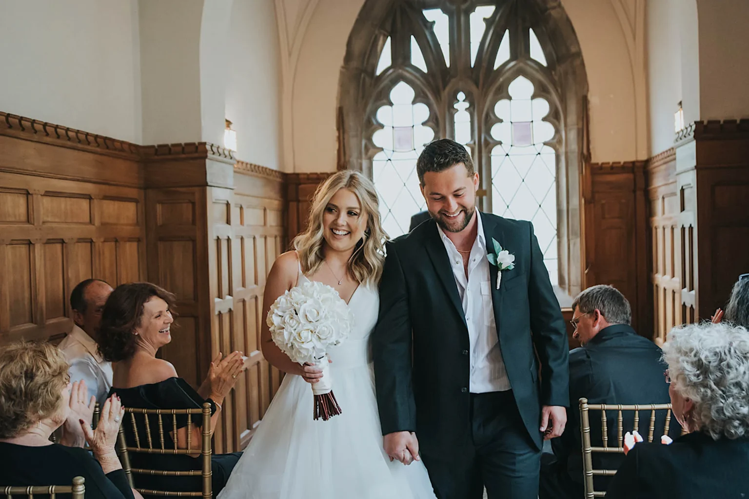 Elopement Wedding A smiling bride in a white gown holding a bouquet and a groom in a dark suit walk down an aisle in a wood-paneled hall with arched windows. Guests seated on both sides, some clapping and others smiling, celebrate their marriage. The atmosphere is light and joyful. Elopements Inc