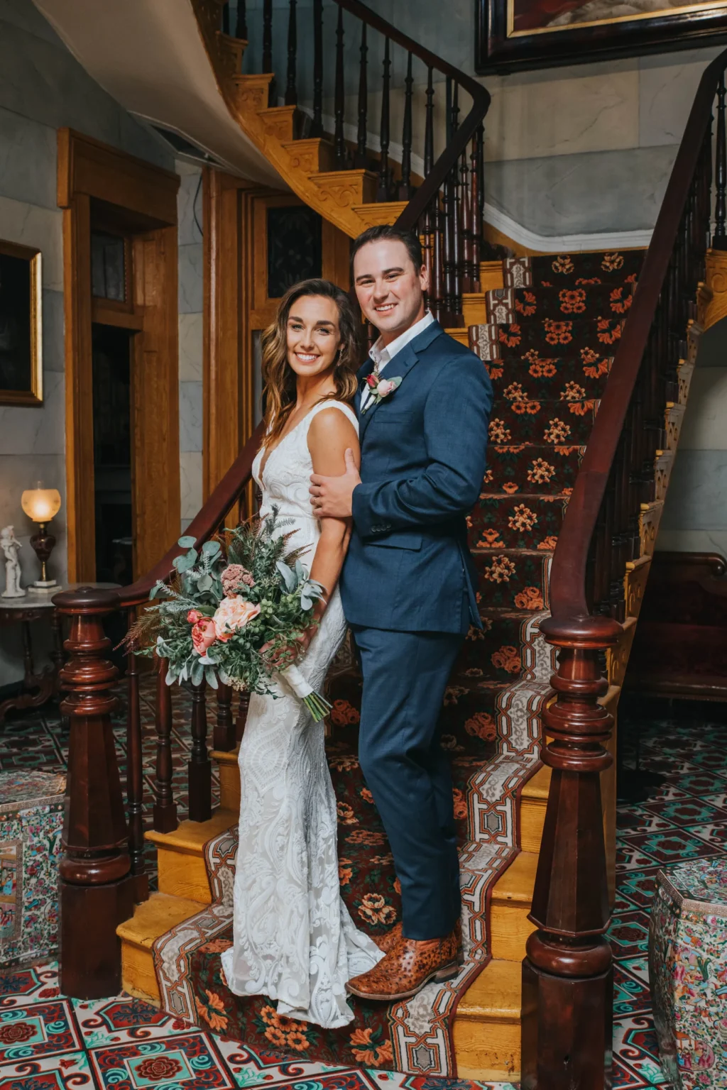 Elopement Wedding A bride and groom smile at the base of a wooden spiral staircase. The bride wears a white lace gown and holds a bouquet of flowers. The groom is in a blue suit. They stand on vibrant patterned tiles and an intricately designed carpet on the stairs. Framed art and elegant decor adorn the background. Elopements Inc