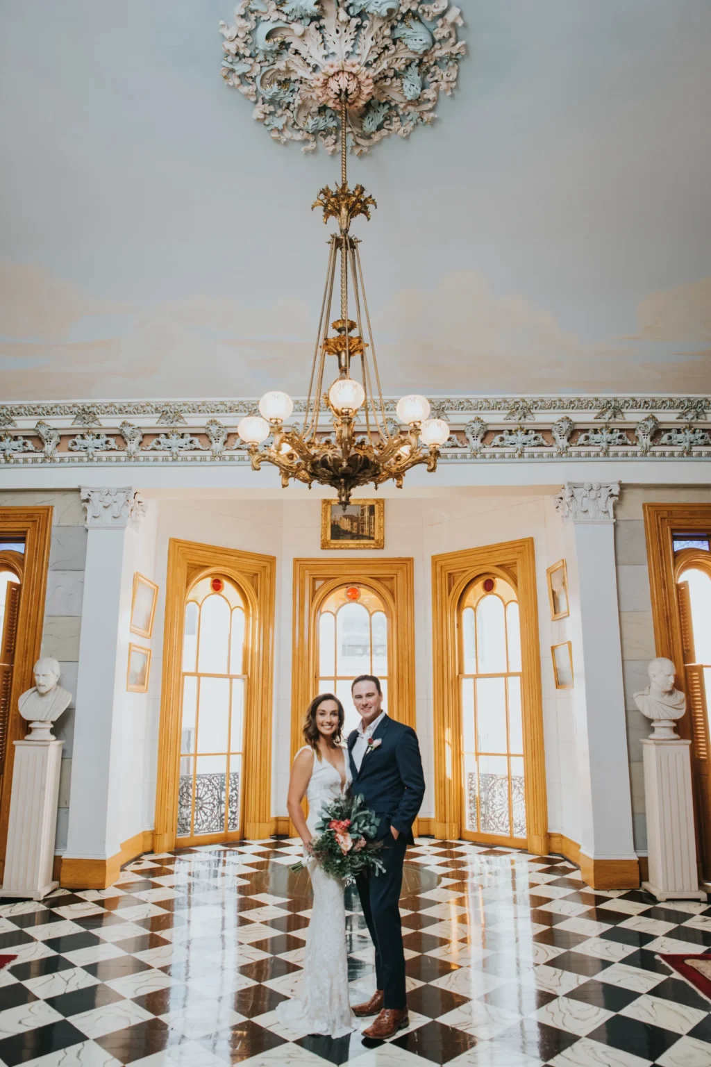 Elopement Wedding A bride in a white gown and groom in a dark suit pose in an ornate hall with high ceilings, large arched windows, and intricate chandeliers. The floor features a black and white checkered pattern, and the walls are adorned with classical statues and framed pictures. They are holding a bouquet of flowers. Elopements Inc