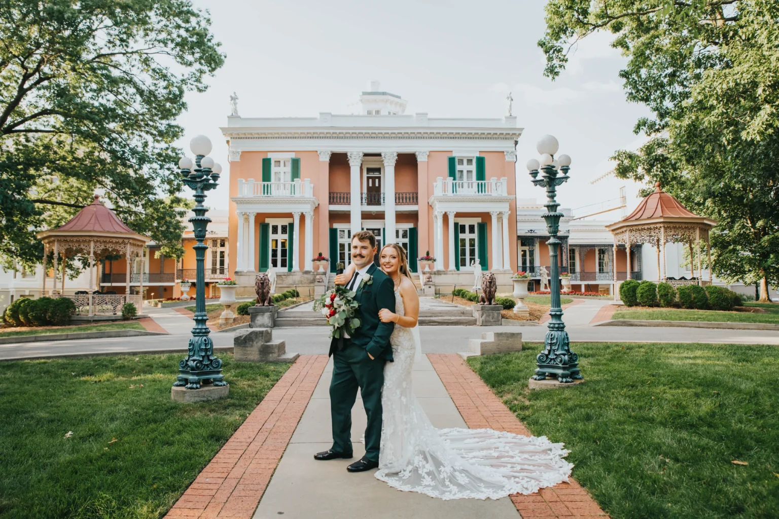 Elopement Wedding A bride and groom stand arm in arm on a walkway in front of an elegant mansion with white columns, balconies, and green shutters. The bride wears a white lace dress with a long train, and the groom is in a dark suit, holding a bouquet. Two gazebos flank the path, and trees surround the scene. Elopements Inc