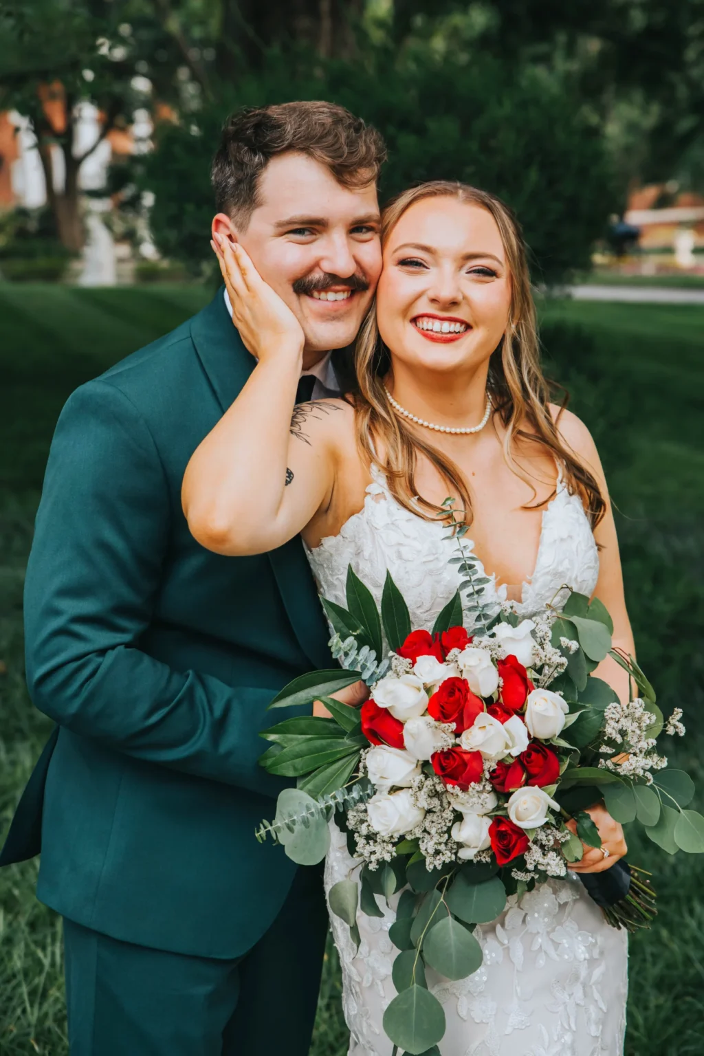 Elopement Wedding A smiling couple dressed in wedding attire poses outdoors. The groom, in a dark green suit, is embraced by the bride, who wears a white gown and holds a bouquet of red and white flowers with greenery. They stand against a backdrop of lush greenery, exuding joy and affection. Elopements Inc