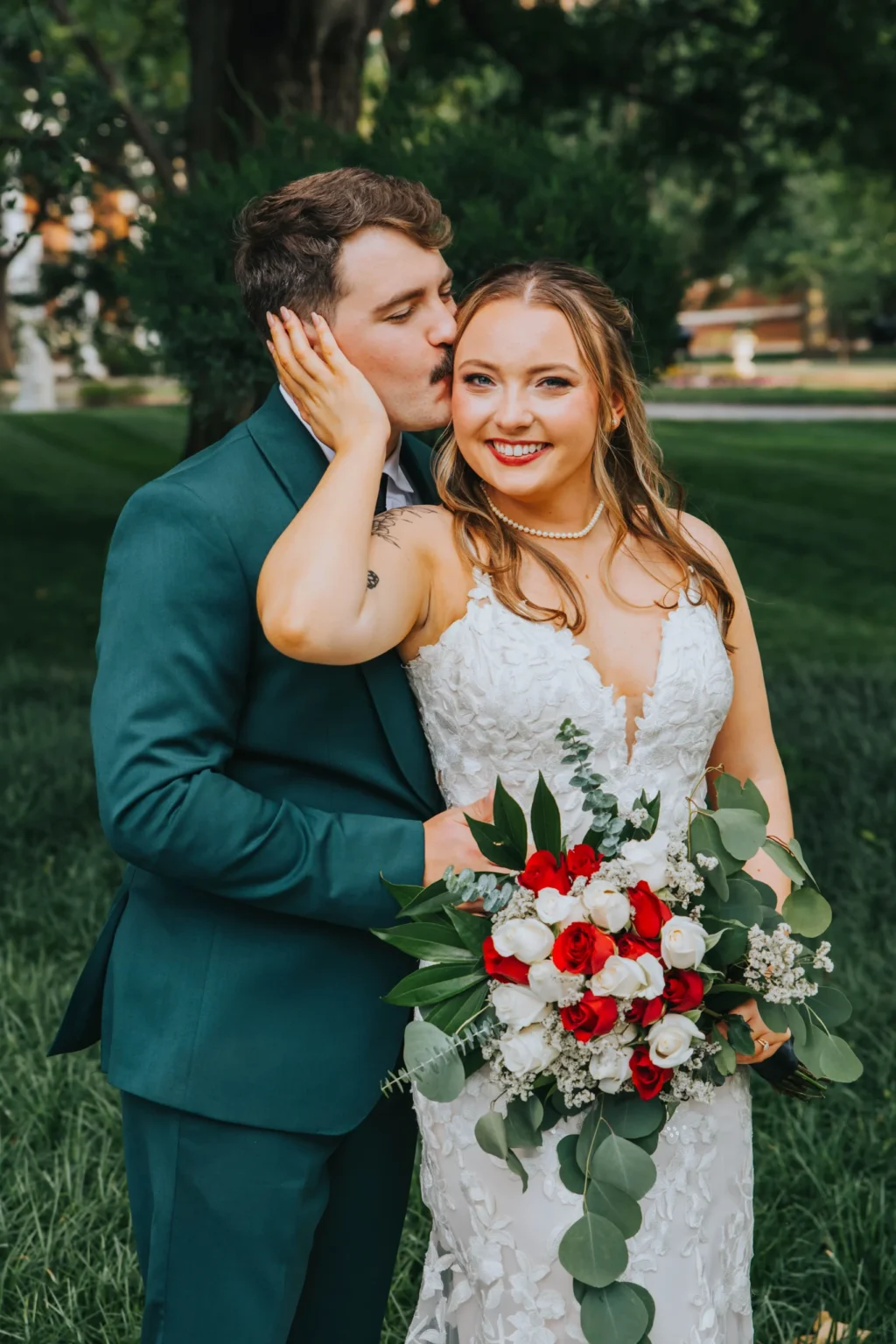 Elopement Wedding A groom in a green suit kisses the cheek of a smiling bride in a white lace gown. The bride holds a bouquet of red and white roses with greenery. They stand in a lush, green outdoor setting with trees and a building in the background, sharing an intimate and joyful moment. Elopements Inc