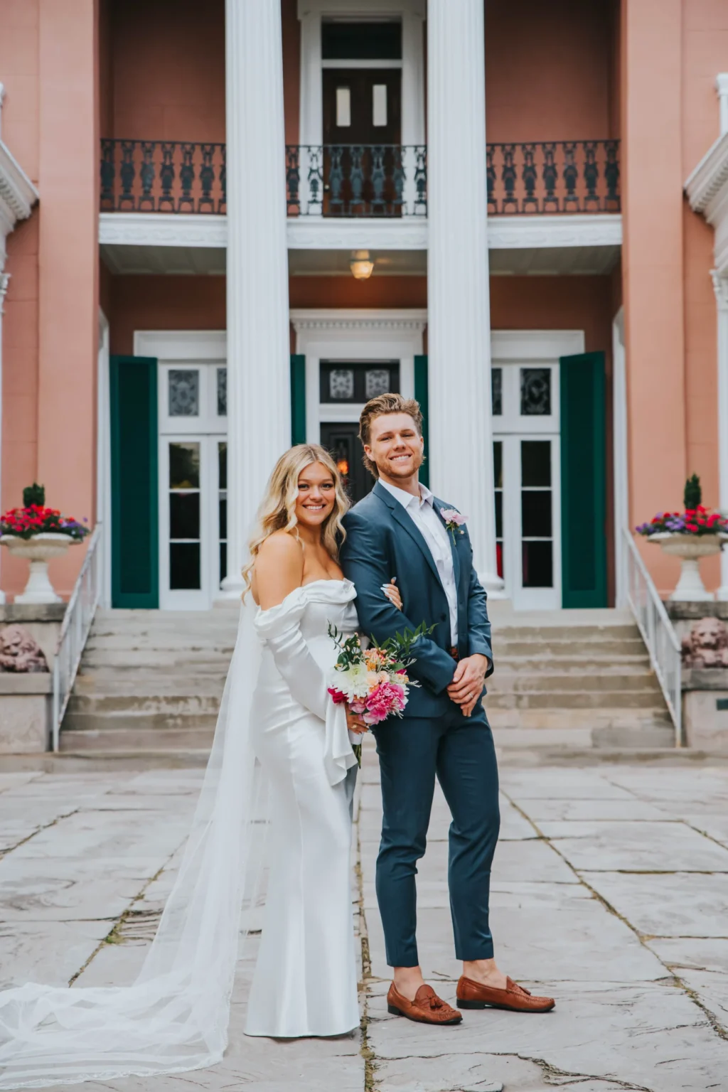 Elopement Wedding A bride in a white off-shoulder gown holding a bouquet and a groom in a blue suit stand arm in arm smiling in front of a building with large white columns and double doors. The building has a reddish exterior, green shutters, and colorful potted flowers on both sides of the entrance. Elopements Inc